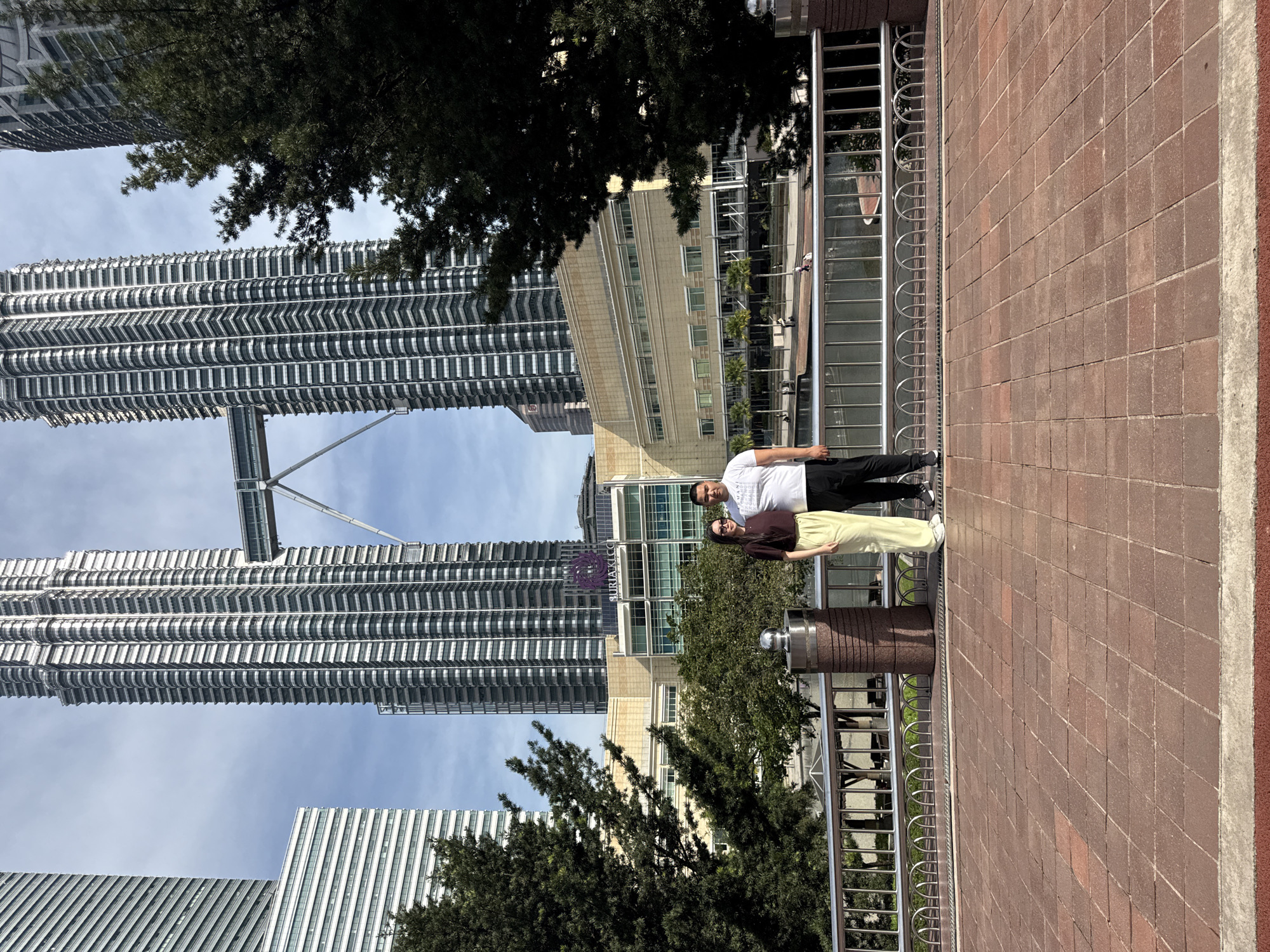 The image shows a couple standing side-by-side on a red-brick pedestrian walkway, with a metal railing behind them. The background features the Petronas Twin Towers in Kuala Lumpur, Malaysia. The couple appears to be posing for a photo. The surrounding environment includes other buildings and trees. The sky is partly cloudy.