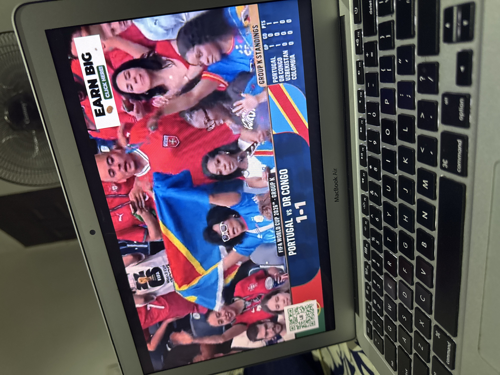The image shows a laptop displaying a soccer match on its screen. The match appears to be between Portugal and DR Congo, with the score currently 1-1. The screen shows a group of spectators in the background, some of whom are holding flags. There is also an advertisement banner at the top of the screen that reads "EARN BIG CLICK HERE". The laptop is open on a surface that appears to be a bed, with a fan visible in the background. The image is taken from a slightly high angle, looking down at the laptop. The location is not specified as Kufena, Nigeria.