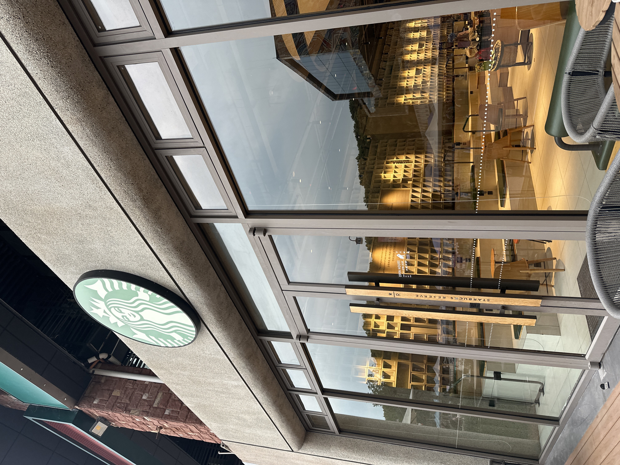 The image captures an exterior view of a Starbucks Reserve store, featuring a modern glass facade with prominent reflections. The photograph is taken from a low, upward angle and is rotated 90 degrees clockwise, presenting the building at an unconventional tilt.
To the left of the frame, a distinct round Starbucks logo, depicting the green and white siren, is mounted on a textured, light-grey concrete wall. Below this section, a band of reddish-brown brickwork is visible.
The majority of the image is dominated by the store's extensive glass window system, framed by dark metal. These windows act as a mirror, reflecting a bright, overcast sky and the surrounding architecture, including a modern building with dark glass and geometric lines, as well as hints of distant greenery.
Through the glass, the interior of the Starbucks Reserve store is clearly visible, illuminated by warm lighting. A defining feature inside is a wall lined with numerous wooden shelving units, each composed of many small, square compartments. Cafe furniture, including wooden tables and chairs, is arranged on a light-toned floor. A small round table near the entrance holds decorative items. To the far right, parts of outdoor seating, characterized by grey woven chairs, suggest an adjacent patio area.
Visible text includes "STARBUCKS RESERVE" prominently displayed on vertical wooden panels that serve as door handles. Above this, a sign reads "OPEN HOURS" followed by Chinese characters, indicating the store's operating times and confirming its location in Chongqing High-tech Zone, China. Fainter English and Chinese text about the Starbucks Reserve concept can also be seen. The scene is set during daytime, with a bright sky reflected, complementing the warm interior lighting.