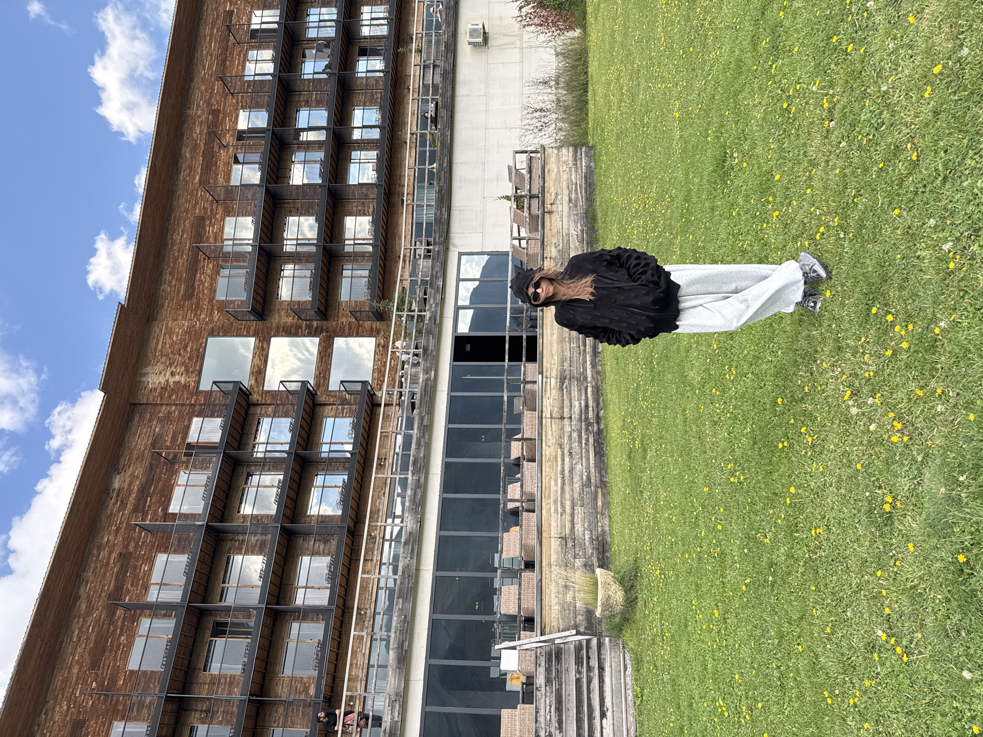 A full-body, eye-level shot shows a young woman standing in a grassy field in front of a large, wooden building. The woman is wearing a black hooded jacket with its hood up, light gray sweatpants, and dark sneakers. She has on dark sunglasses with a heart-shaped frame and her hands are in her pockets. The building behind her is multi-storied with numerous balconies and windows. A set of stairs leads up to a patio area with wicker furniture. The grass in the foreground is green and dotted with yellow dandelions. The sky above is blue with scattered white clouds. The location is identified as Stepantsminda, Georgia, suggesting the building is likely a hotel or resort.