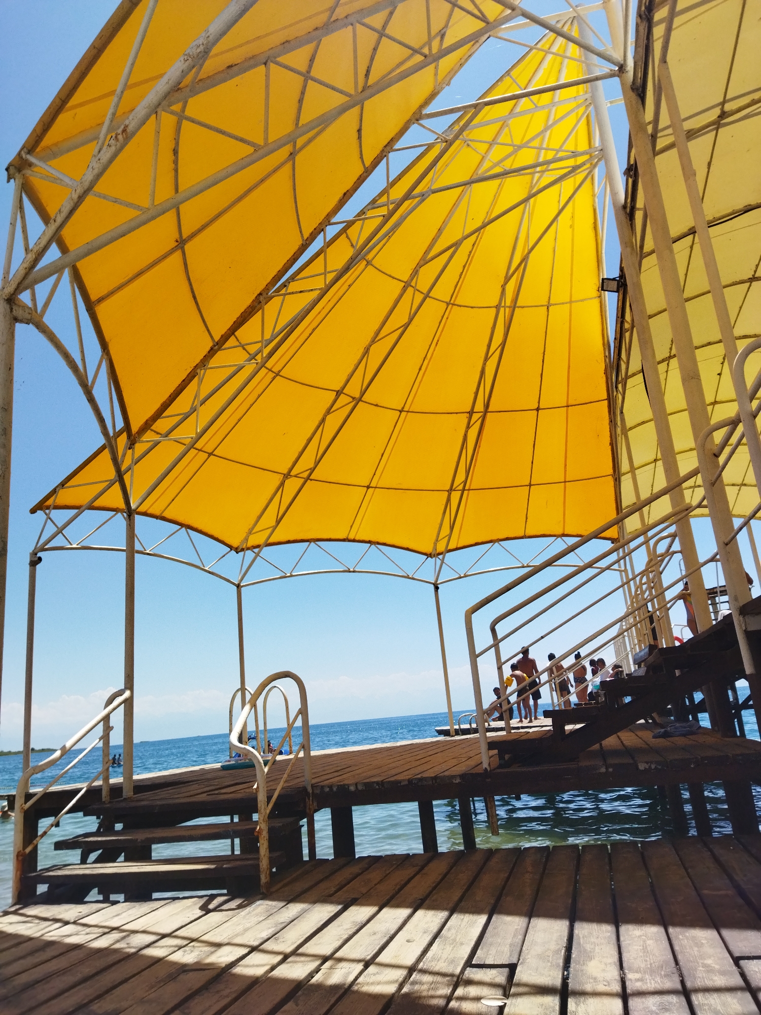 The image depicts a sunny day scene at a beach or lakefront area in Kara-Oy, Kyrgyzstan. In the foreground, a wooden pier with weathered planks leads towards a body of water. Steps are visible on the pier, with white metal railings. Overhead, large yellow canvas awnings supported by a white metal frame provide shade. These awnings are taut and appear to be designed to catch the wind, resembling sails.
Beyond the pier, a calm blue body of water stretches to the horizon, with a faint shoreline visible in the distance. The sky is clear and bright blue.
Several people are present on the pier and in the water. On the pier, individuals are seen walking up and down the stairs and standing near the edge. In the water, a few people are swimming or wading. One person appears to be standing near a small raft or inflatable device.
The overall impression is one of a recreational area, likely a resort or public beach, during daylight hours. The scene is bathed in bright sunlight, creating strong shadows on the wooden pier. The yellow awnings are a prominent visual element, adding a splash of vibrant color against the blue sky and water.