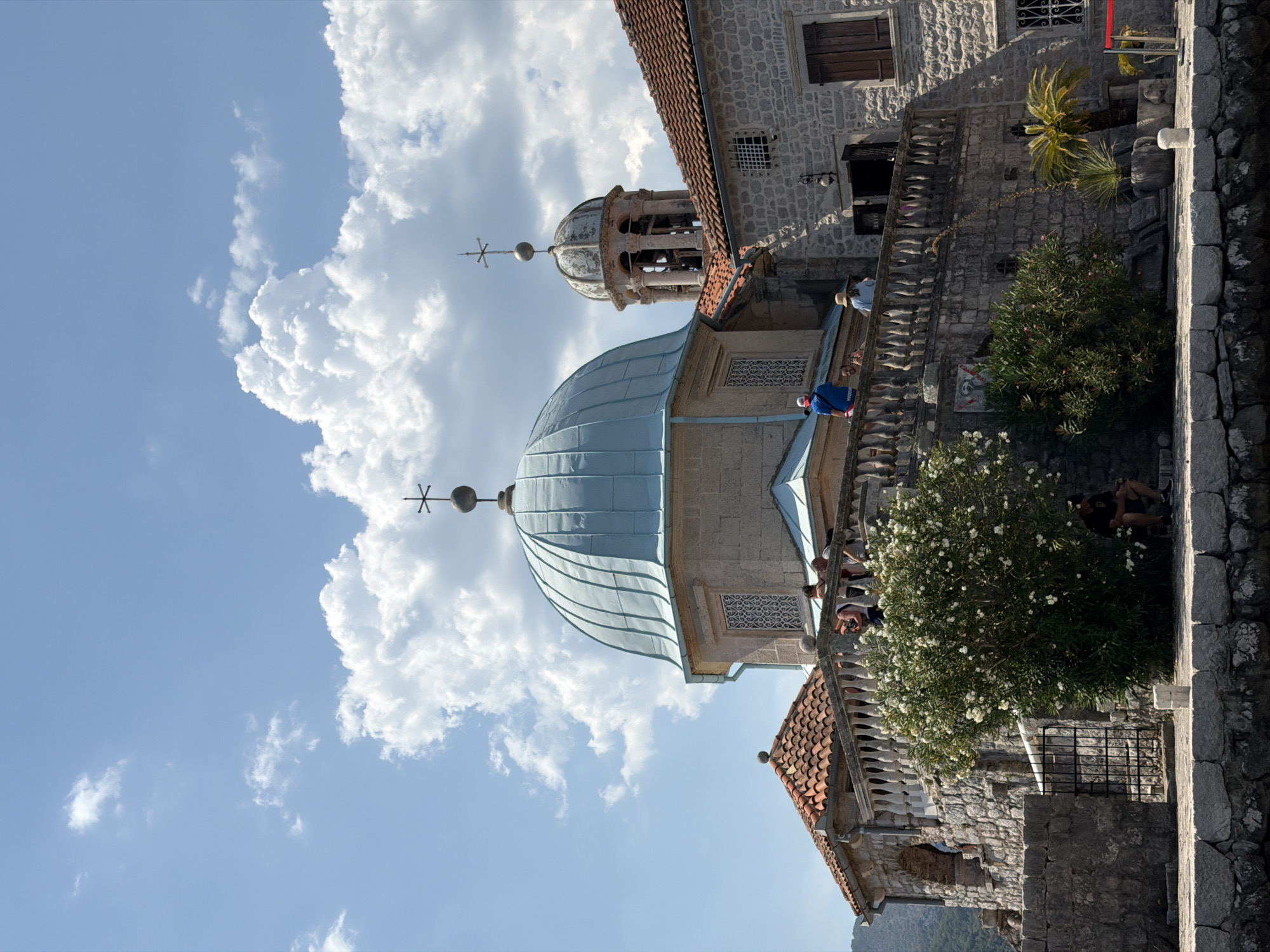 This is a vertical, eye-level, outdoor, daytime photograph taken of the Church of Our Lady of the Rocks, an island church located in Perast, Montenegro. The church has a prominent light blue domed roof with a smaller bell tower with a dome behind it. The buildings are constructed of light-colored stone with red-tiled roofs. A stone balcony overlooks the water, with several people standing on it, observing the surroundings. There are lush green bushes and trees in the foreground, and a man is sitting on a bench on the right side of the frame. The sky is a clear blue with scattered white clouds. The water is calm and reflects the sky.