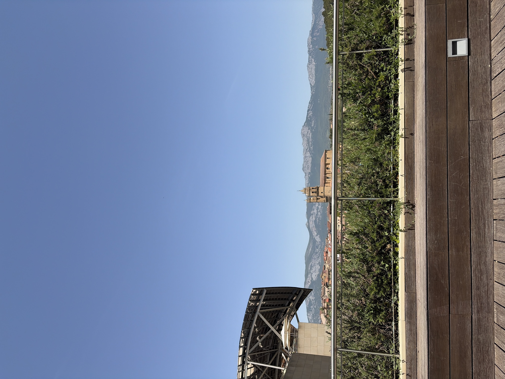 The image shows a view from a rooftop with wooden decking, looking out over the city of Unknown, Unknown. A metal railing separates the viewing area from greenery and the city below. The skyline is dominated by mountains in the background. In the middle ground, a church tower and other buildings are visible. A modern structure with an arched roof is in the lower left foreground. The sky is clear and blue.
