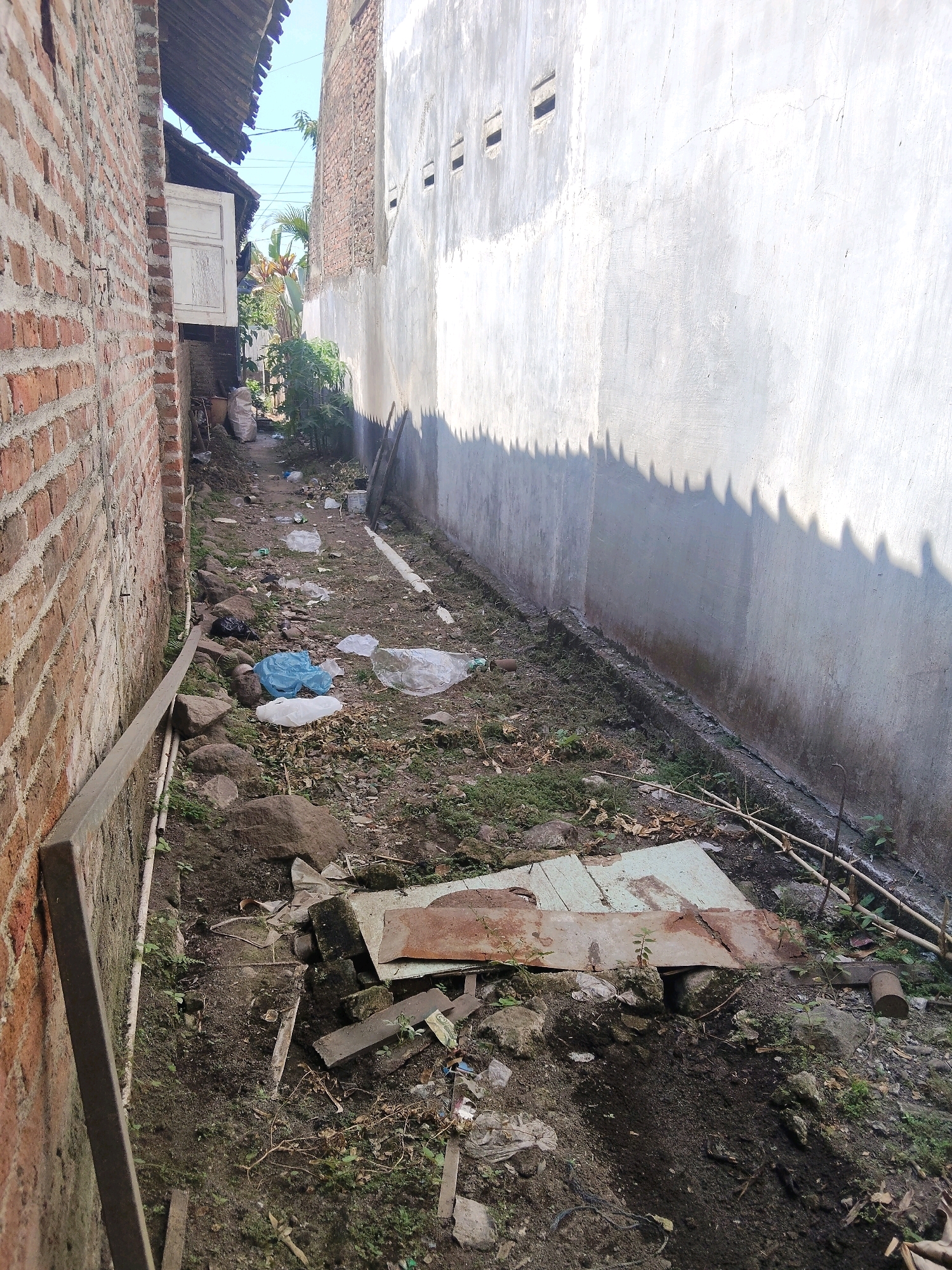 The image depicts an outdoor scene in Windurejo, Indonesia, likely a narrow alleyway or side yard between two buildings. A brick wall is visible on the left, while a light-colored, textured concrete wall runs along the right side. The ground is unpaved, with patches of dirt, grass, and debris. Scattered across the ground are various discarded items, including plastic bags, rocks, and pieces of wood or metal. The overall impression is one of a neglected or utilitarian space.