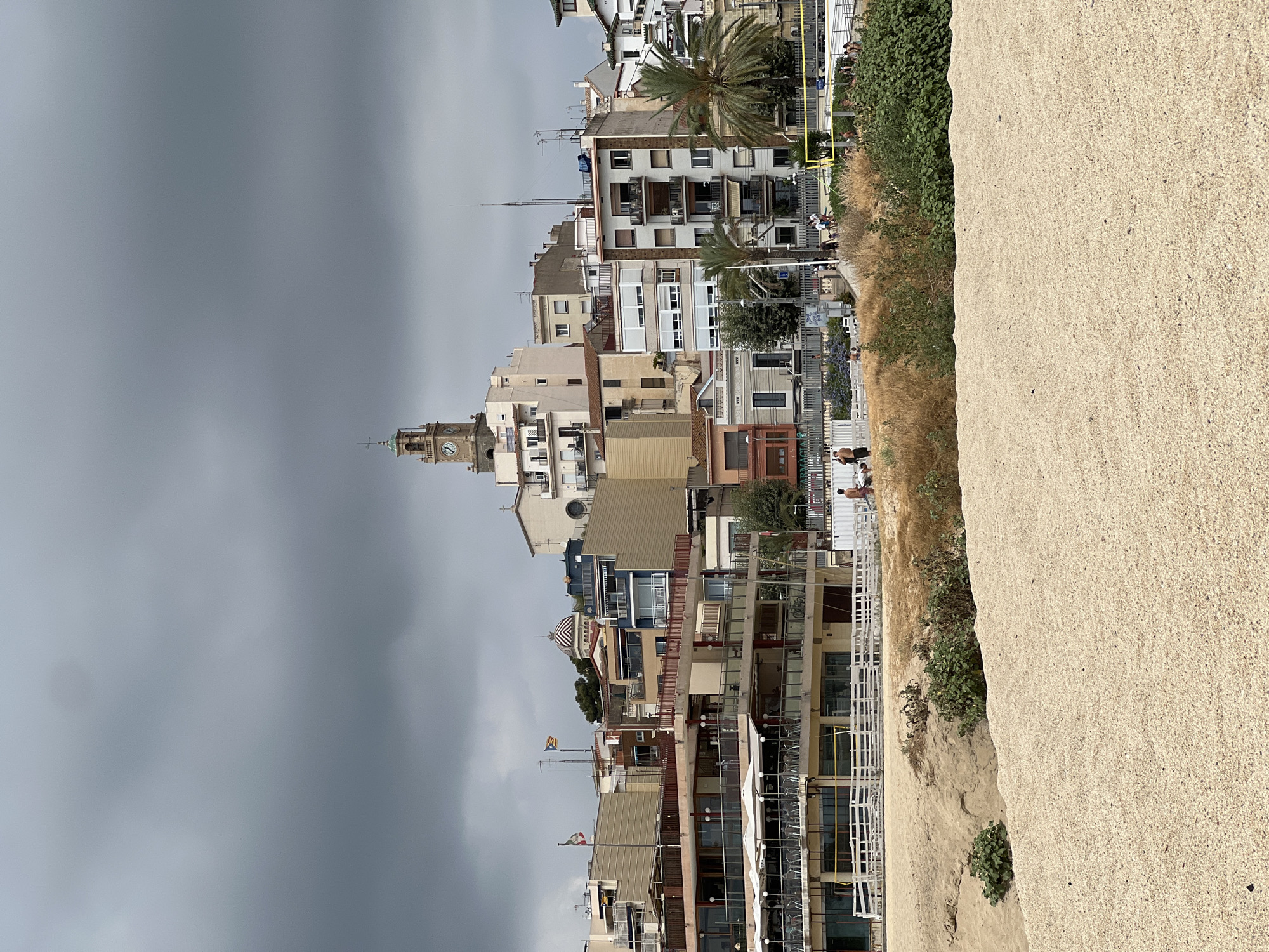 The image shows a view of the town of El Masnou, Spain, from a sandy beach. In the foreground, there is a large expanse of light-colored sand with some sparse vegetation. Behind the beach, there is a building with multiple levels, likely a restaurant or beach club, with outdoor seating and umbrellas visible. Further back, the town rises up a hill, characterized by a dense cluster of buildings of varying heights and architectural styles. A prominent clock tower, painted beige with a green-topped spire, stands out above the other structures. Several flags are visible on poles atop some of the buildings. The sky is overcast with dark gray clouds, suggesting potentially inclement weather. A few people are visible on the beach and near the buildings, some walking and others appearing to be relaxing. The overall scene depicts a coastal town with a beach resort area.