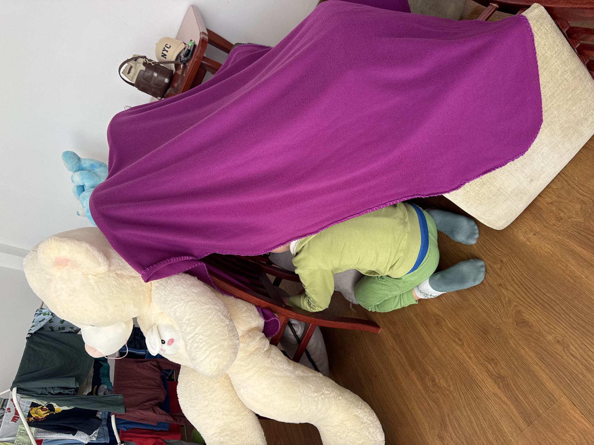 A child wearing a green pajama set and blue socks is partially visible under a purple blanket draped over a cushioned stool. A large teddy bear is positioned near the stool, partially under the blanket. The scene is set on a wooden floor, and a clothes rack with folded clothes is visible in the background. A white wall is also visible in the background. The location is not indicated in the image, there is no indication this photo was taken in Burgas, Bulgaria.