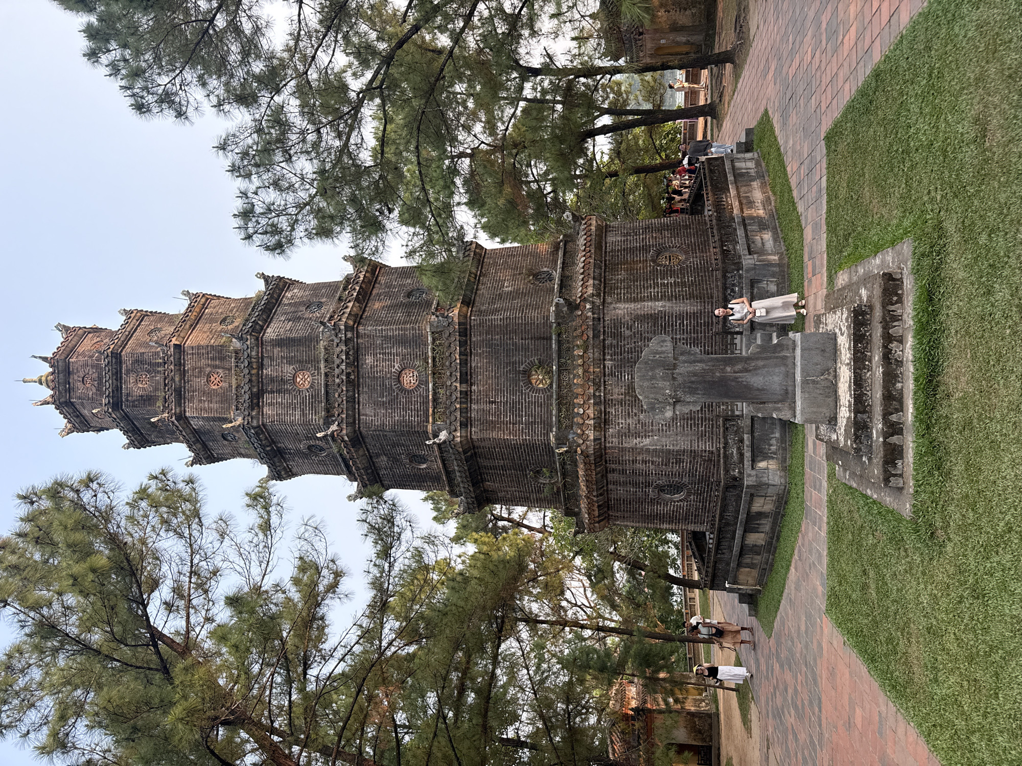 The image depicts the Thien Mu Pagoda in Kim Long Ward, Vietnam, a tall, multi-tiered structure built of dark brick. The pagoda is surrounded by trees. A woman in a light-colored dress stands on a stone structure near the pagoda's base. People are visible further back, near a stone building. The ground is a mix of brick paths and green grass.