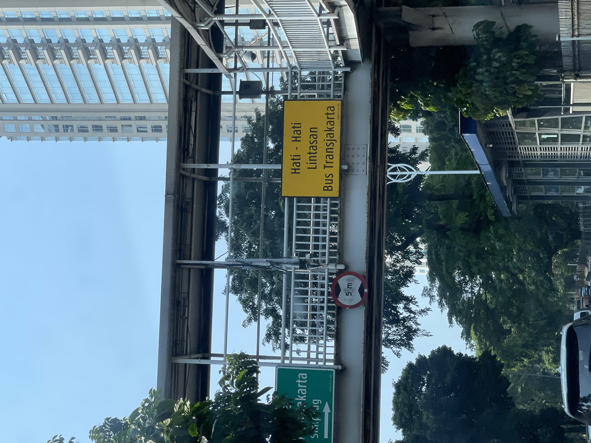 The image shows a street-level view of an elevated pedestrian walkway or overpass in South Jakarta, Indonesia. The overpass is constructed with a metal frame and has a covered roof. On the walkway, there are several signs. One green sign displays text in white, including "Jakarta" and "Simpug" with a directional arrow pointing to the right. Adjacent to this is a circular red and white sign with "5 m" inside, indicating a height restriction. Another prominent sign is rectangular and yellow, featuring text in Indonesian: "Hati - Hati Lintasan Bus Transjakarta," which translates to "Be Careful Transjakarta Bus Crossing." The overpass overlooks a busy road with vehicles, including cars and buses, and is lined with lush green trees. In the background, a modern, tall building with glass windows stands against a clear blue sky. Below the overpass, a bus stop structure with glass panels is visible. The overall scene depicts an urban environment with public transportation infrastructure and traffic.