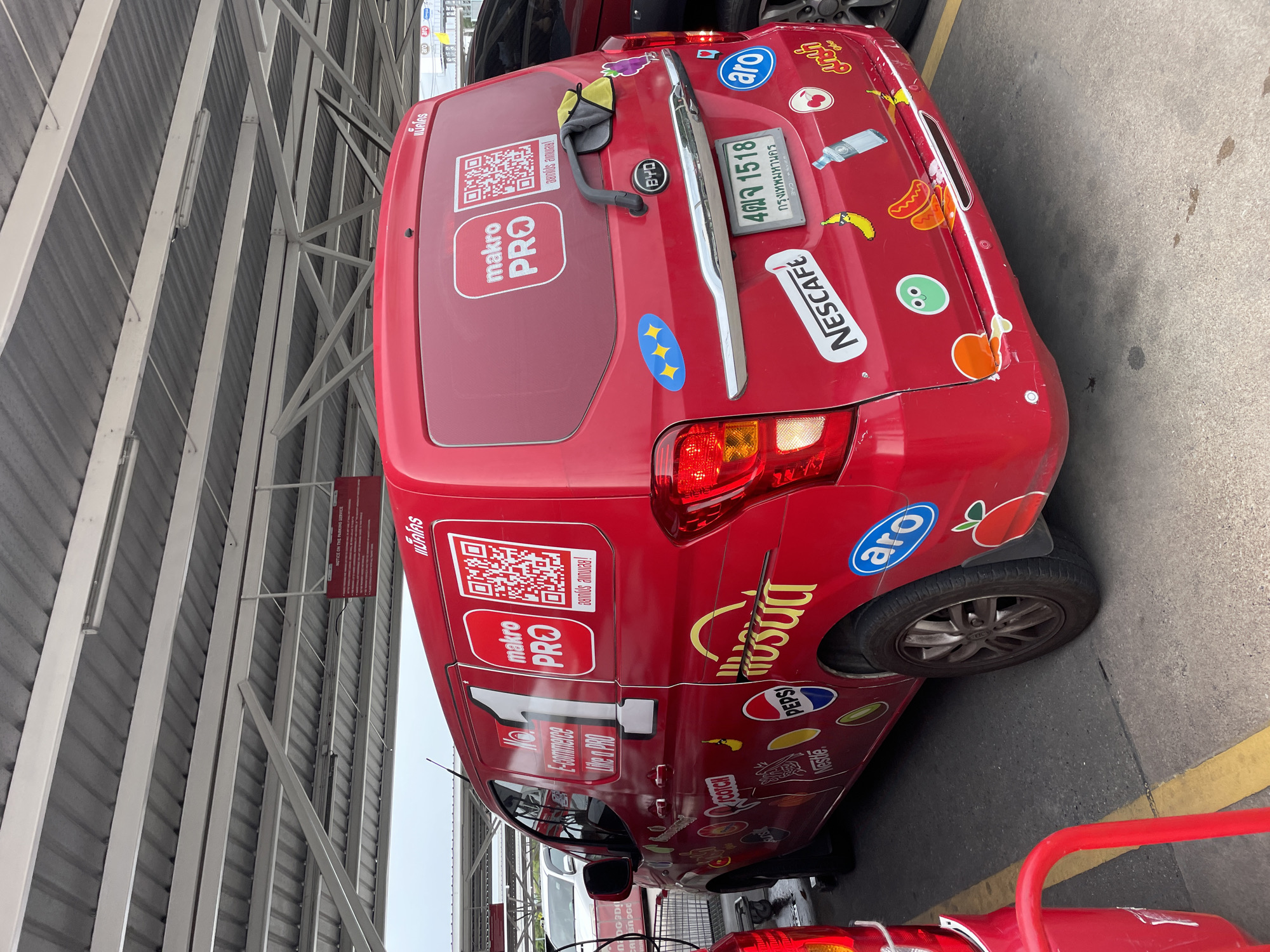 The image shows the rear and side of a red delivery van, likely a BYD, parked under a covered parking structure. The van is extensively decorated with stickers, including logos for "Makro PRO," "Nescafe," "Pepsi," and "Nestle," as well as cartoonish images of food items like hot dogs and bananas, and a green cartoon face. A license plate with Thai characters and the number "4พจ 1518" is visible on the rear of the van. In the background, another vehicle is partially visible, and a red shopping cart is to the left of the van. The environment suggests a commercial or retail area, consistent with a location in Pattaya City, Thailand, given the Thai script on the van and license plate. The covered structure appears to be made of metal beams and corrugated roofing.