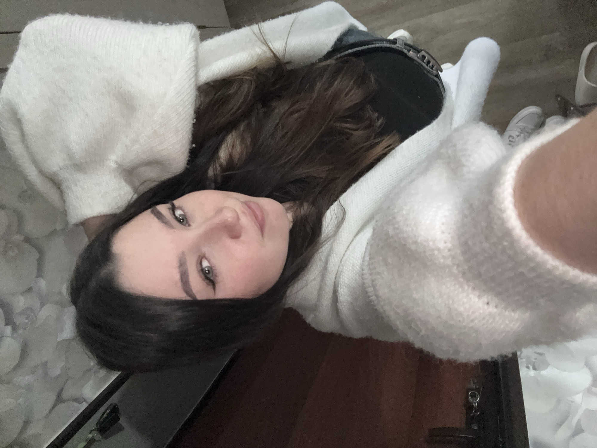 A selfie of a young woman with dark brown hair, wearing a white cardigan over a black top. She is lying down, looking up at the camera with a slight smile. Behind her, there is a glimpse of a light-colored floor and white shoes. Part of a dark brown surface (possibly wood) and a dark object (likely a table or frame) are visible in the foreground. The photograph was taken indoors, likely in a residential setting. The location of Perm, Russia is not discernible from the image.