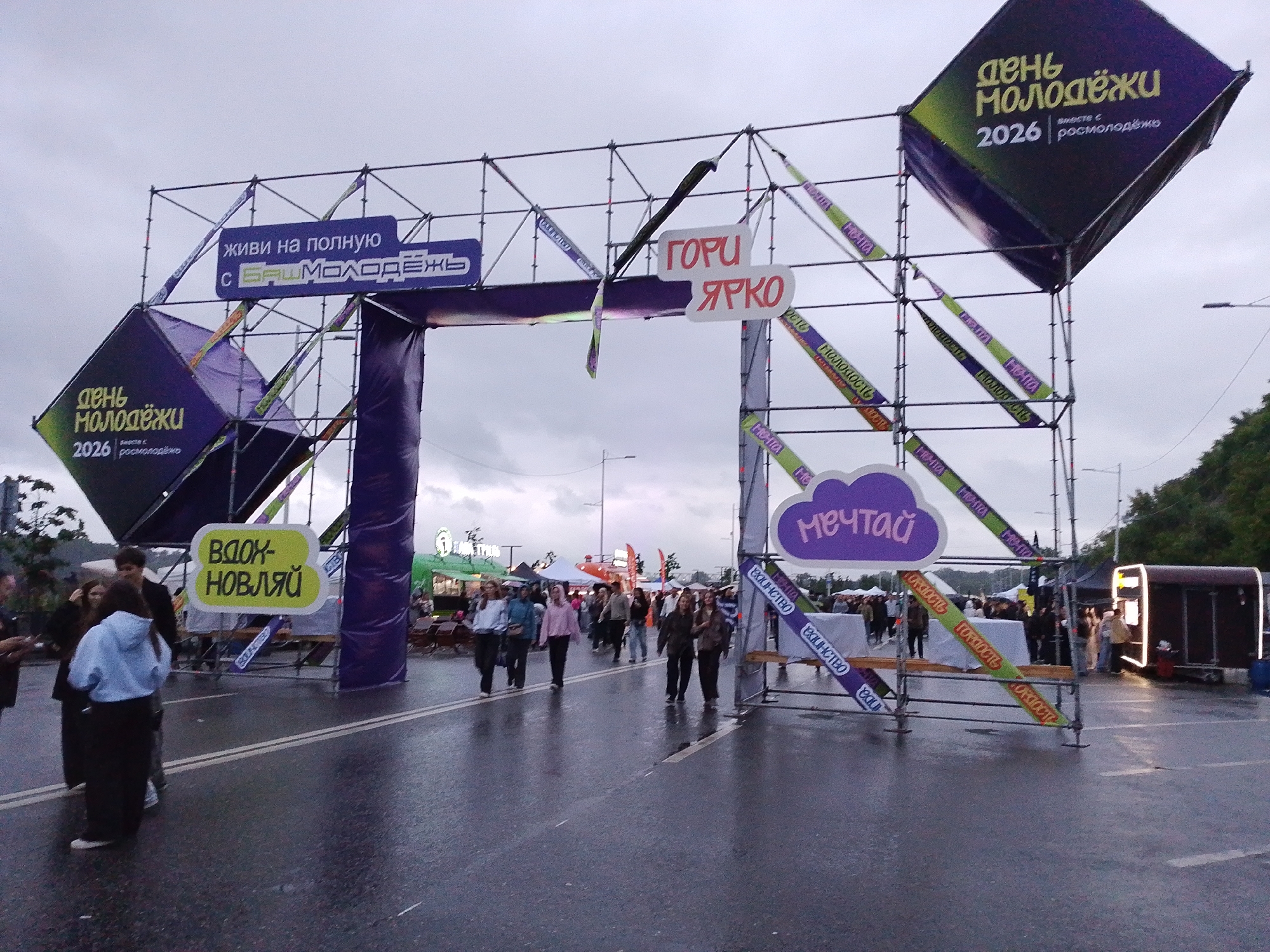 The image depicts an outdoor event, likely a festival or celebration, with a large, arch-like structure serving as an entrance or stage. The structure is adorned with banners and large, cube-shaped decorative elements. On the left side of the arch, a purple cube features text that reads "День молодежи 2026" and "с БАШМОЛОДЕЖЬ". A blue banner above it says "живи на полную с БАШМОЛОДЕЖЬ". On the right side, a purple cloud-shaped sign reads "Мечтай". Various ribbons with repeating text are attached to the structure. In the foreground, people are walking on a wet asphalt surface, suggesting it may have rained or is raining. Various stalls or tents are visible in the background, along with more people. The scene suggests a lively event taking place in a public space. The text "БАШМОЛОДЕЖЬ" strongly suggests the event is related to Bashkortostan, a republic within Russia, and Ufa is its capital city.