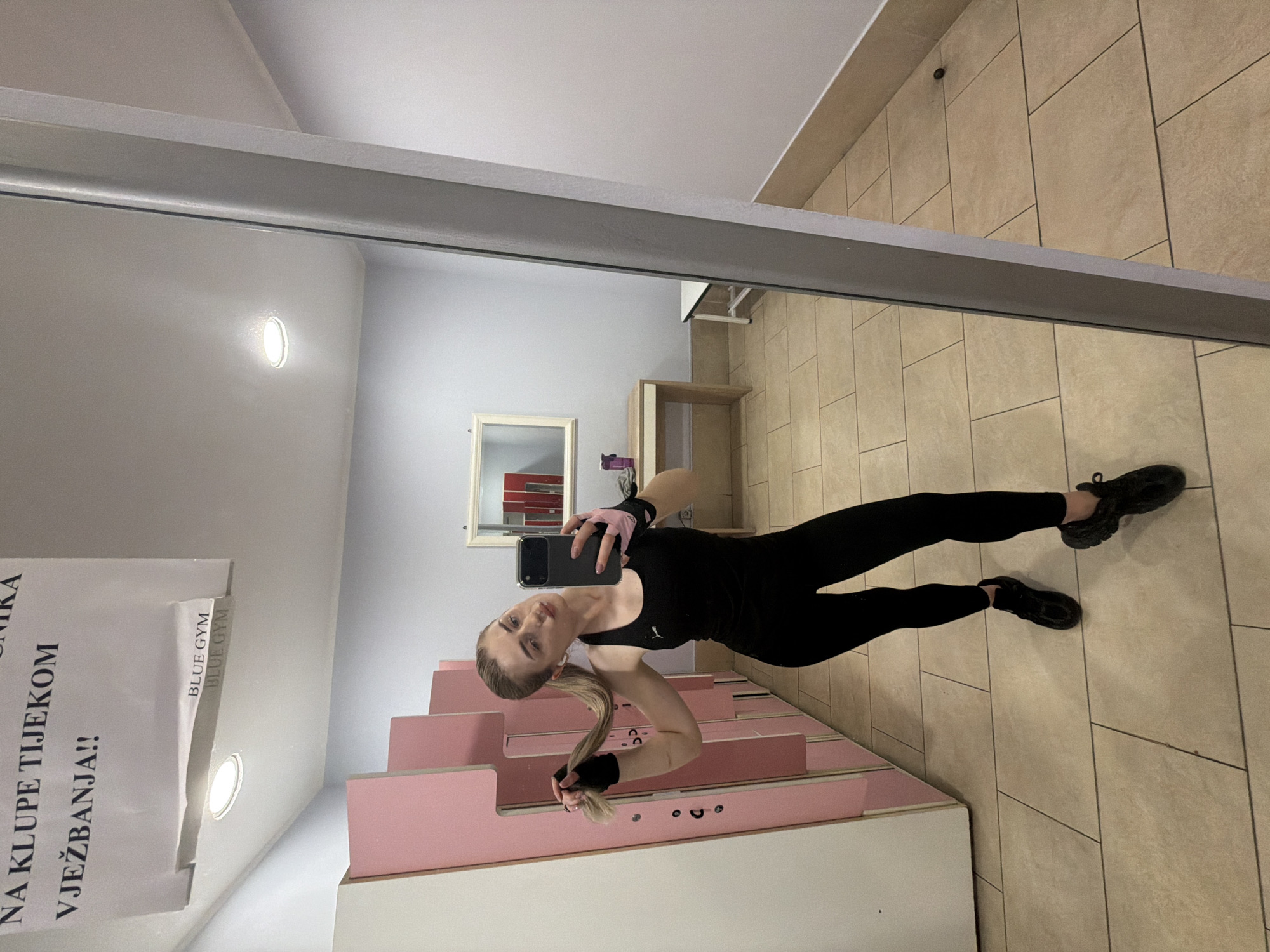 A young woman with blonde hair tied in a ponytail, wearing a black athletic tank top and leggings, takes a mirror selfie in a gym locker room. She is holding her ponytail with her left hand, which is covered by a black wristband, and her right hand is holding a smartphone. She is also wearing black athletic shoes and pink fingerless gloves. The locker room has light pink lockers to her left and a long mirror to her right. In the background, there is a desk with a mirror above it, and a small white bench. The floor is tiled in a light beige color. A sign is partially visible on the ceiling above the lockers with text in a foreign language.