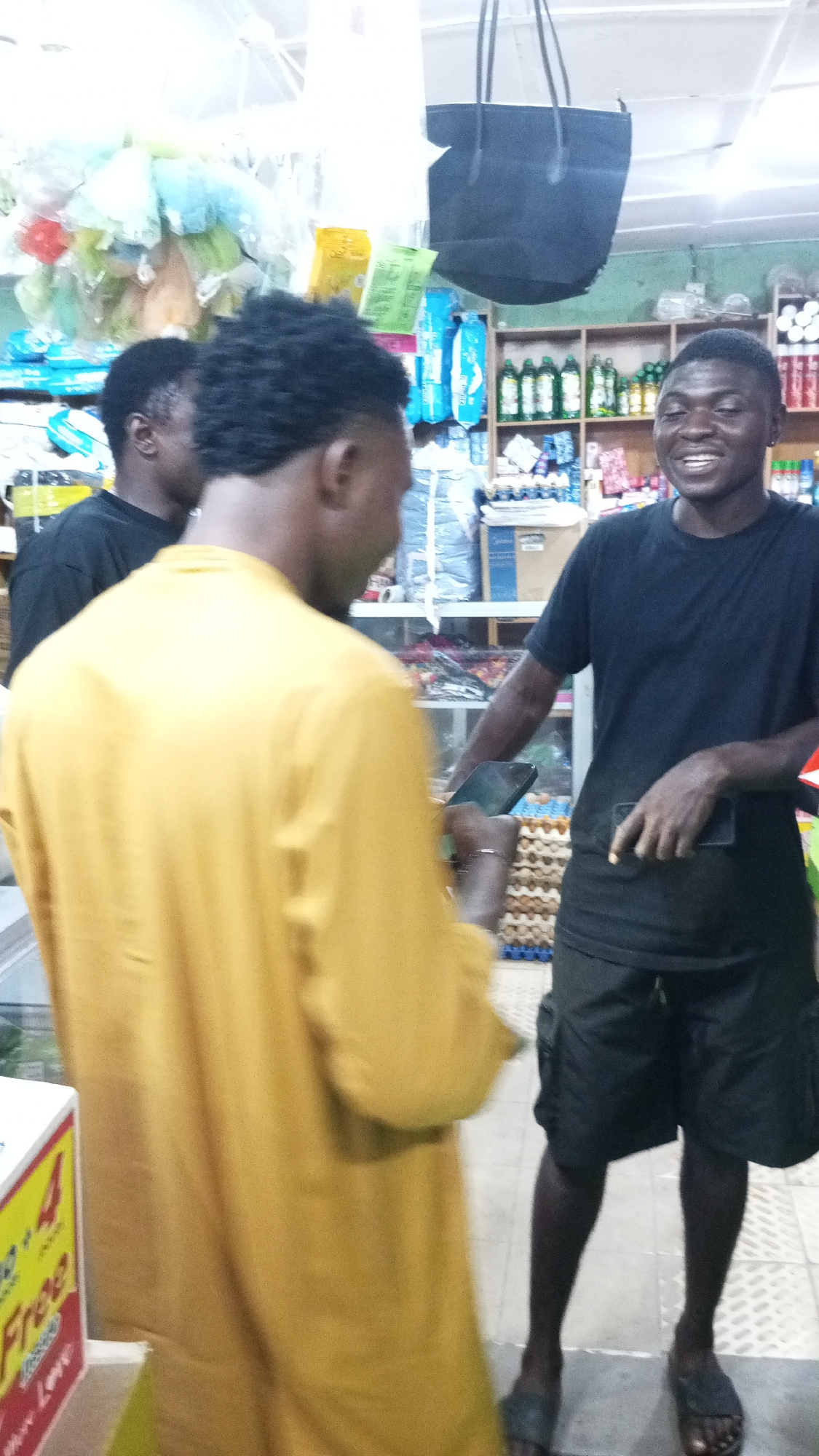 The image depicts an indoor scene within what appears to be a local convenience store or market. Three men are visible. In the foreground, with his back mostly to the camera, is a man wearing a long-sleeved mustard yellow shirt and dark, curly hair. He is turned towards the right side of the frame, seemingly interacting with another man.
The second man, positioned on the right and in clearer focus, is smiling broadly. He is wearing a black t-shirt, black shorts, and black slides. He holds a black smartphone in his left hand, and possibly another small black object or phone in his right hand. His gaze and smile are directed towards the man in the yellow shirt, indicating a friendly exchange or conversation. A third man is partially visible behind the man in the yellow shirt, wearing a black t-shirt.
The background reveals typical store shelving stocked with various products, including rows of green bottles, packaged goods, and stacks of eggs in blue and brown crates on the floor. A black tote bag hangs from the ceiling in the upper center, and numerous light-colored plastic bags are also suspended overhead. On the far left, a white and yellow sign with red text is partially visible, clearly stating "Buy 4 Get 1 Free" and "Free" underneath, suggesting a store promotion. The setting is brightly lit, consistent with artificial indoor lighting. The overall impression is a casual interaction between the individuals within a retail environment.