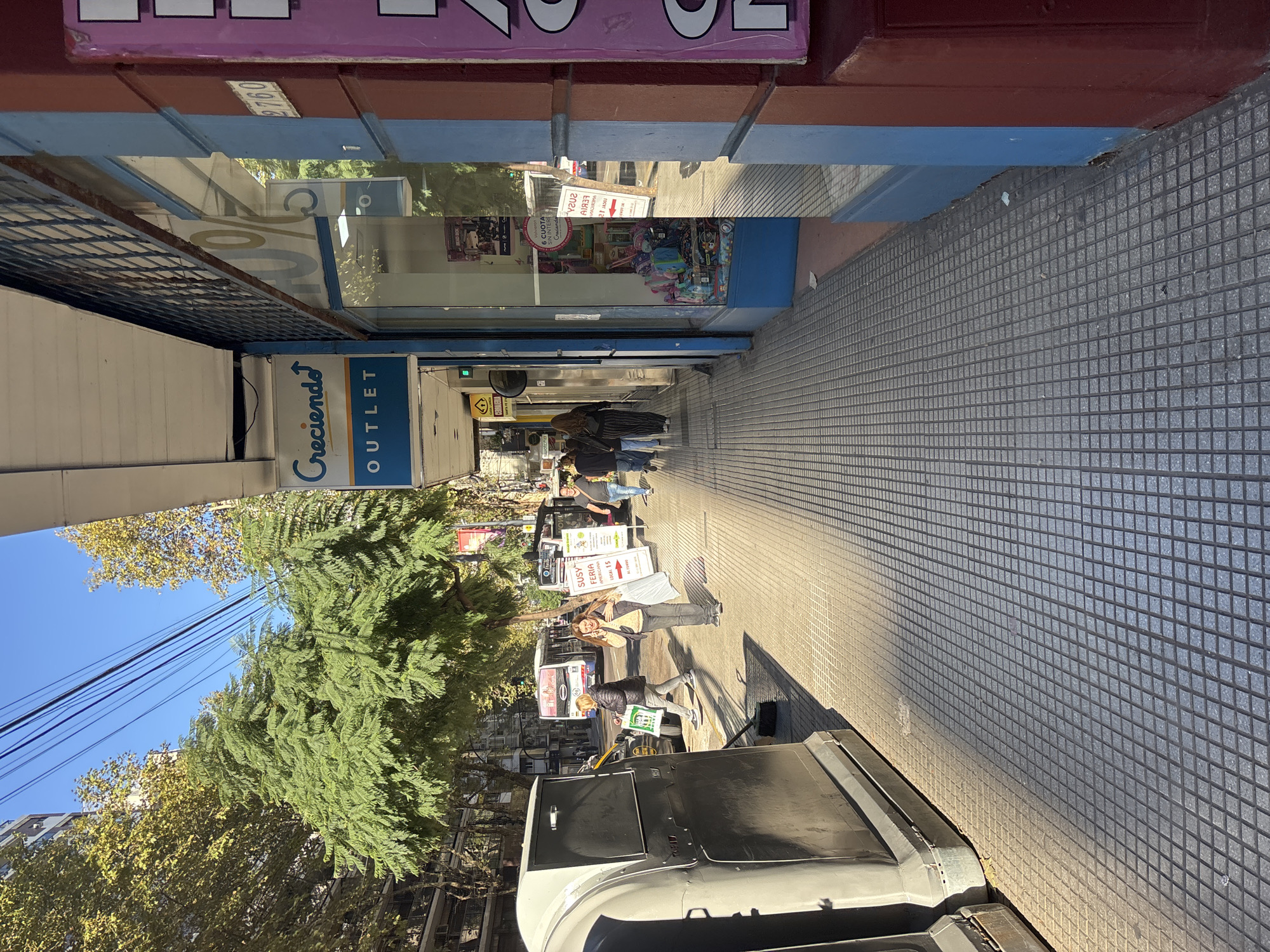 This image captures a vibrant urban street scene, likely in Buenos Aires, Argentina, on a clear, sunny day. The perspective is looking down a sidewalk lined with shops, featuring a mix of pedestrians and street activity.
On the left side of the frame, several storefronts are visible. Prominently displayed is a blue and yellow sign for "Creciendo! OUTLET," a children's goods store. Further down, other shop windows show merchandise and signs, including a partially visible "CUOTA SIN INTERES" (installments without interest) indicating payment options. A building number "9760" is seen on a red facade.
The sidewalk, made of distinctive grey square tiles, stretches into the distance on the right, bathed in bright sunlight that creates sharp shadows. People are seen walking along the sidewalk; one individual in a dark jacket walks away from the camera, holding a green item, while another person stands near a white sign. Further down, additional pedestrians contribute to the lively atmosphere.
Trees with green foliage line the street on the left, casting dappled shadows. Overhead power lines are visible against the bright blue sky. In the immediate foreground, a large, dark-colored vehicle, possibly an SUV or van, is partially visible, parked on the street.
A white freestanding sign on the sidewalk reads "SUSY FERIA AMERICANA" with "LOCAL 15" and "AL FERRA" partially visible, suggesting a second-hand market or fair booth. The overall impression is one of everyday commerce and foot traffic in a bustling city environment.