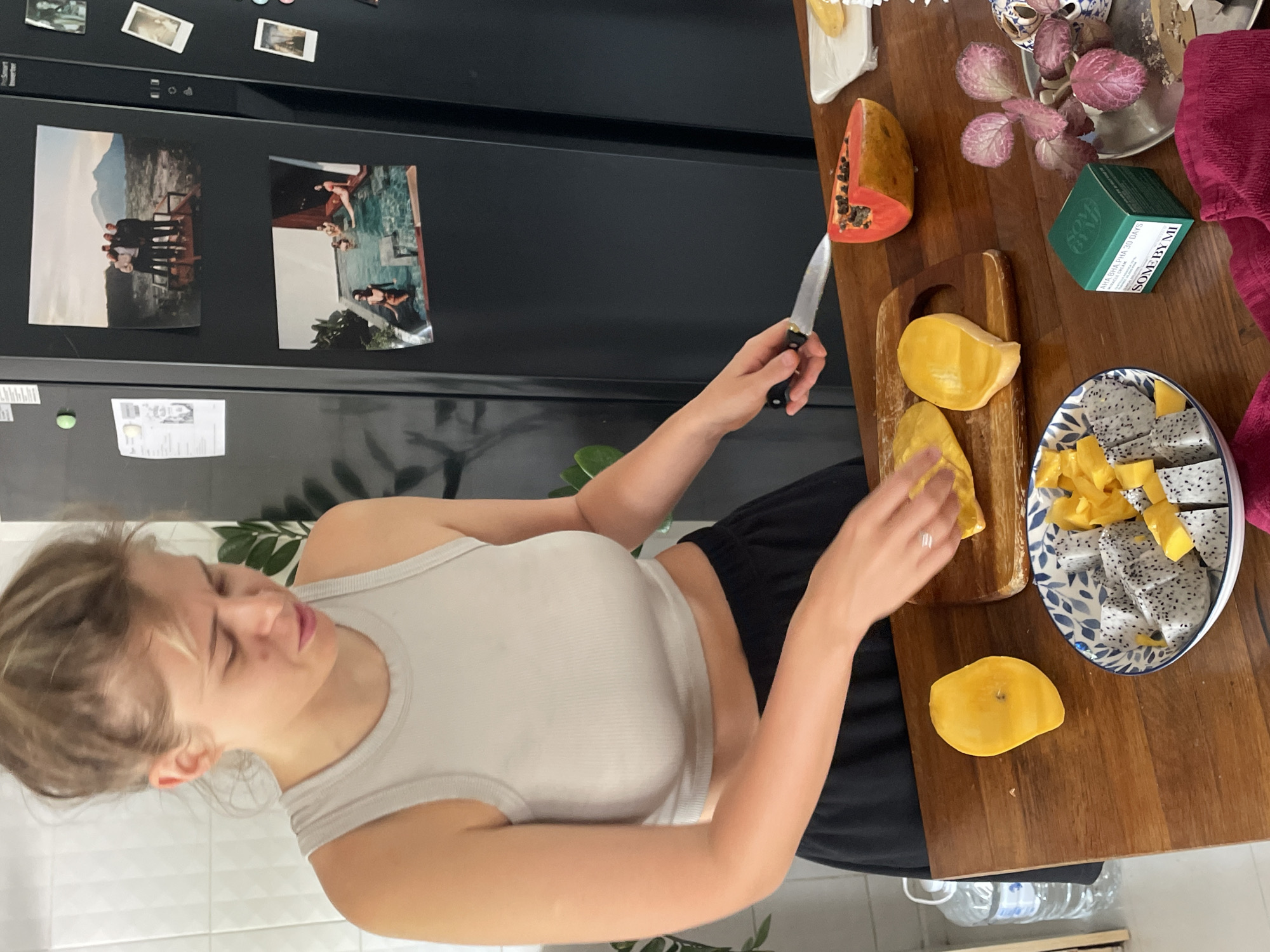 A woman in a white tank top and black shorts is preparing fruit on a wooden table next to a black refrigerator. She is using a knife to cut a mango on a wooden cutting board. Other fruits on the table include papaya and dragon fruit, the latter arranged on a blue and white plate. A green box of SOME BY MI cosmetic product, a plant in a pot, and a red towel are also on the table. Photographs are attached to the refrigerator. The location is likely indoors in Chang Phueak, Thailand.
