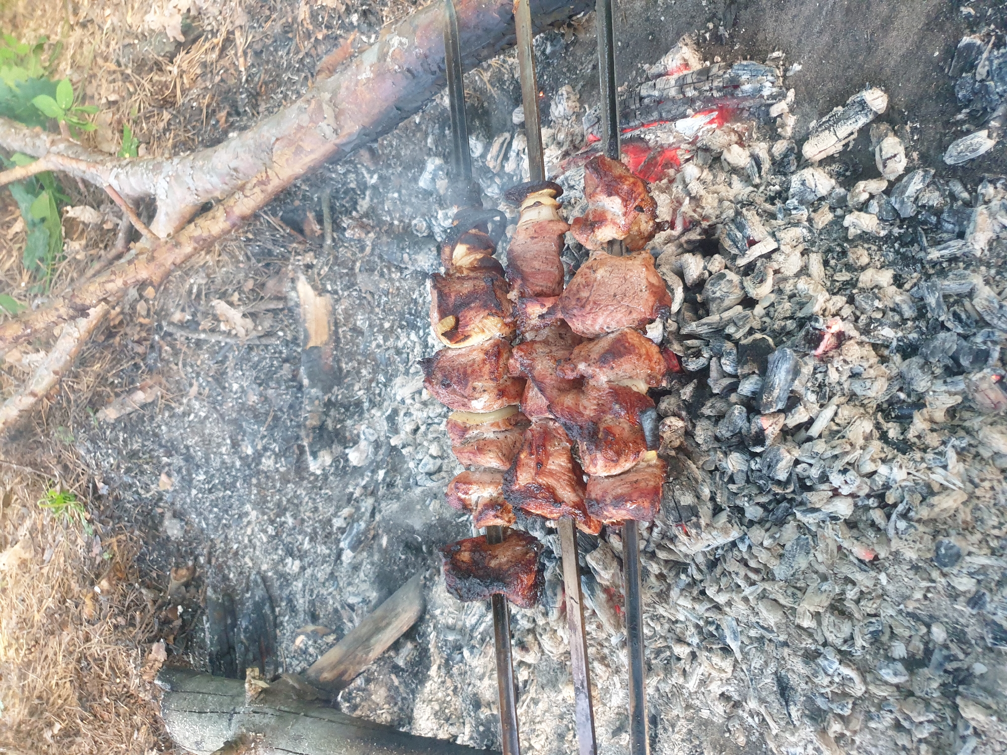 The image shows skewers of meat cooking over coals and embers. The meat appears to be marinated and grilled, with some pieces charred. The skewers are made of metal and are arranged in a row over the fire. The background consists of ash, burnt wood, and dried leaves, suggesting an outdoor cooking setting. There are no people visible in the image. The lighting is natural, and the image is taken from a slightly elevated angle, focusing on the food.
