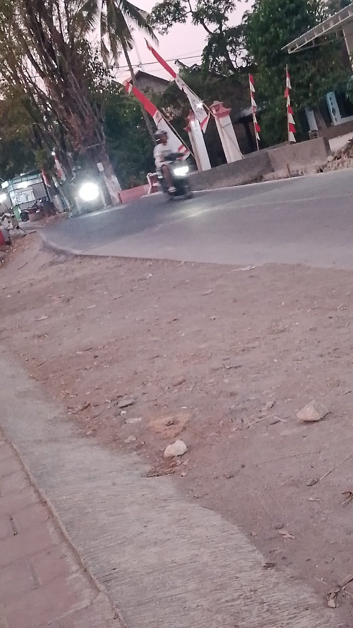 The image depicts a street scene in Karanglangu, Indonesia, at dusk or dawn. A person is riding a motorcycle on a paved road. The motorcycle has its headlights on. On the left side of the road, there are some businesses or stalls with bright lights. Behind the road, there are trees and what appear to be buildings. Several red and white flags, commonly seen during Indonesian independence day celebrations, are displayed on poles. The road curves to the right. The foreground consists of a dirt and gravel area next to a concrete sidewalk.