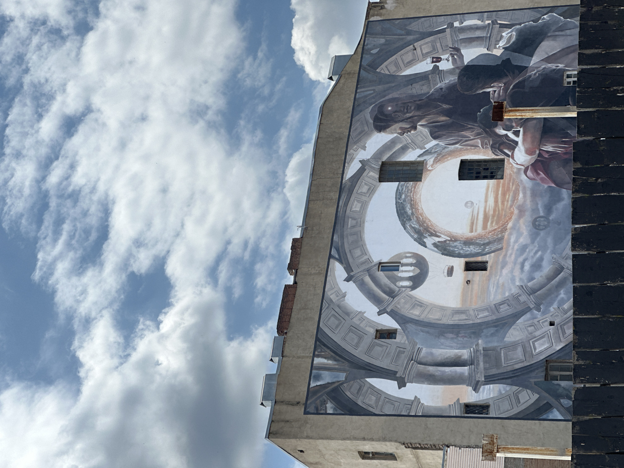 This vertical photograph showcases a striking, large-scale mural adorning the side of a multi-story building in Tbilisi, Georgia, set against a bright, cloudy blue sky. The image is captured from a low angle, emphasizing the impressive height of the building and the artwork.
The mural itself is an intricate, illusionistic piece featuring classical architectural elements and allegorical figures. Prominently positioned towards the upper-center is a figure, possibly a woman, with dark hair and a contemplative expression, holding a stemmed glass containing a reddish liquid. Another face, with luminous detailing near the eye, is visible behind and slightly below the primary figure, integrated into the mural's design.
These figures are enveloped within a complex framework of arches, columns, and coffered structures, rendered in various shades of grey, blue, and brown. These elements create an impressive sense of depth and a grand, dome-like or celestial interior space. The architectural forms appear to curve and rotate, contributing to a dynamic, almost surreal perspective. Towards the bottom of the mural, a large, reflective sphere or globe is depicted, reflecting warm, ethereal clouds or a distant horizon, with smaller spherical elements interspersed throughout.
The building's concrete facade serves as the canvas, with actual windows of varying sizes cut into the wall, subtly integrated into the painted architectural elements. To the right, a dark, heavily textured section of the building's edge, possibly a fire escape or structural support, contrasts with the painted surface. The sky, visible to the left and above the building, is filled with soft, white cumulus clouds against a clear blue background, indicating daytime. No discernible text is visible on the mural or the building. The overall impression conveyed is one of artistic grandeur, skillfully transforming an urban wall into a fantastical and profound scene.
