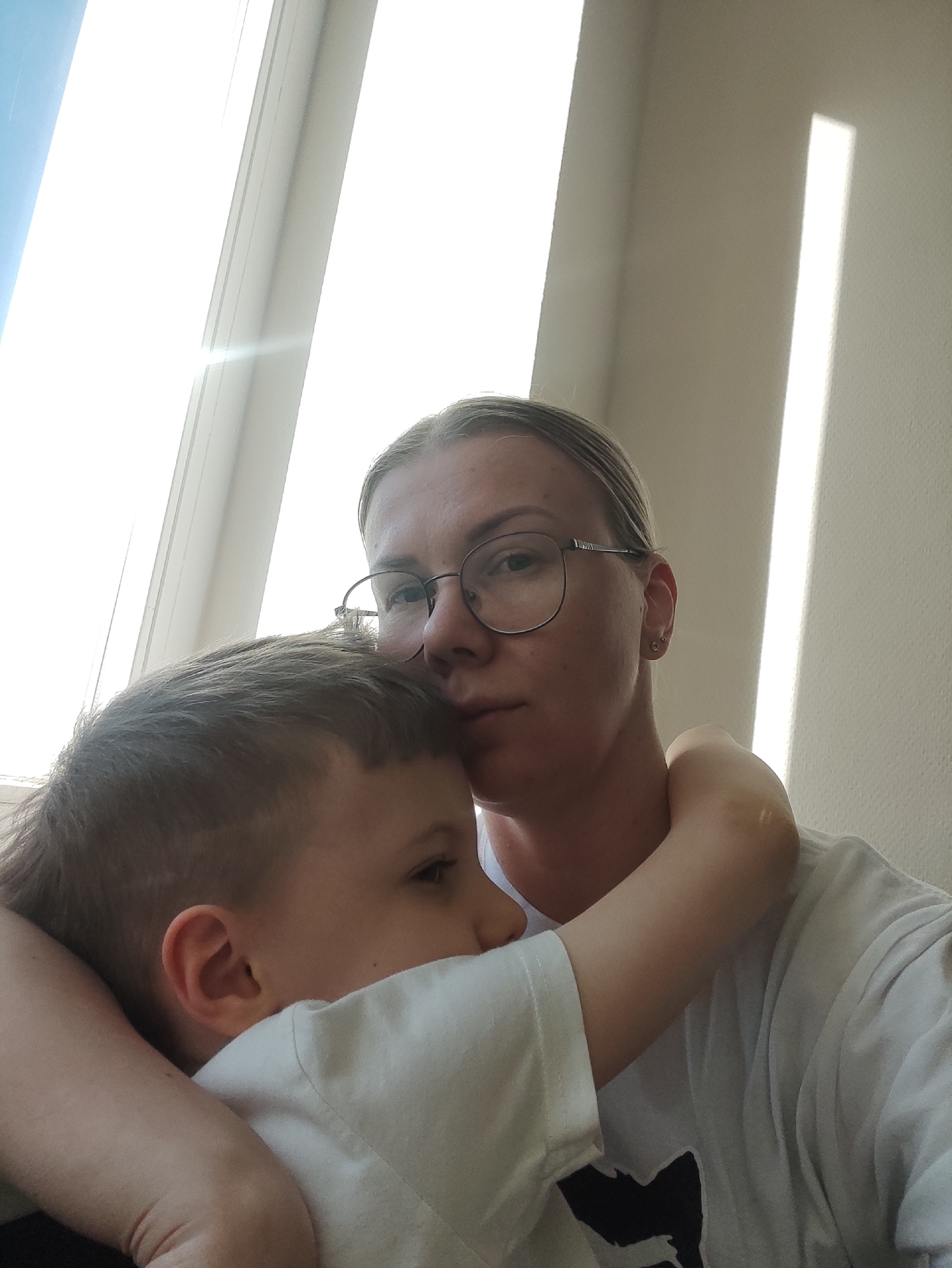 A woman with light skin, fair hair pulled back, and wearing glasses is embracing a young boy with short brown hair. The woman is wearing a white t-shirt. The boy is wearing a light-colored t-shirt. The setting appears to be indoors, possibly near a window, as evidenced by the bright light coming through the window. The background is a plain wall, with a vertical stripe of light visible. The subjects appear to be in Chaikovsky, Russia.