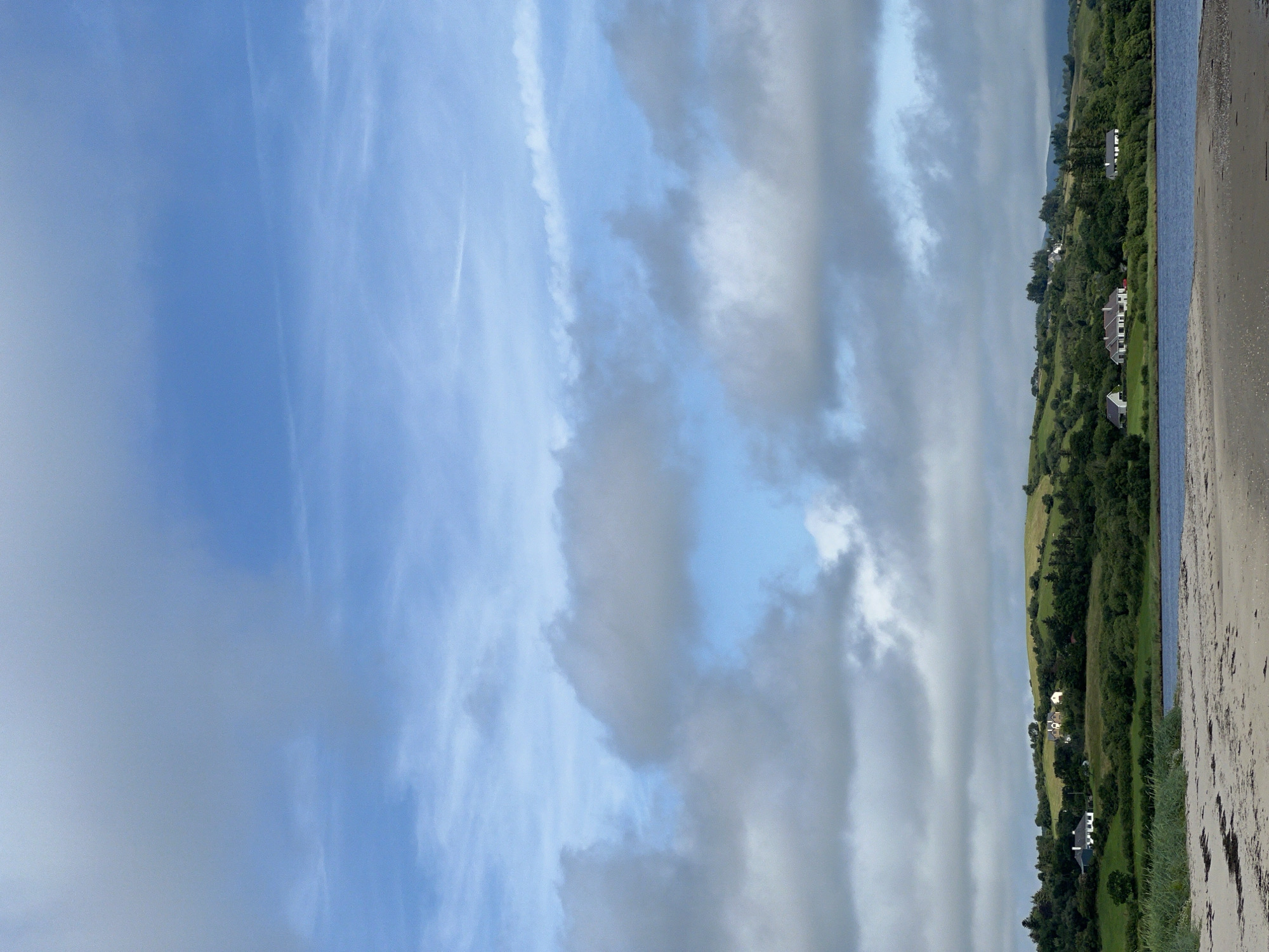 This image depicts a serene landscape scene. In the foreground, a sandy beach with some scattered seaweed leads to a calm body of water. Beyond the water, a lush green hillside is dotted with several houses, some of which appear to be modern residences with white walls and dark roofs. The hillside is covered in a mix of trees and open fields, suggesting a rural or semi-rural setting. The sky above is a pale blue, with scattered, wispy clouds that create a soft, diffused light. The overall impression is one of tranquility and natural beauty.