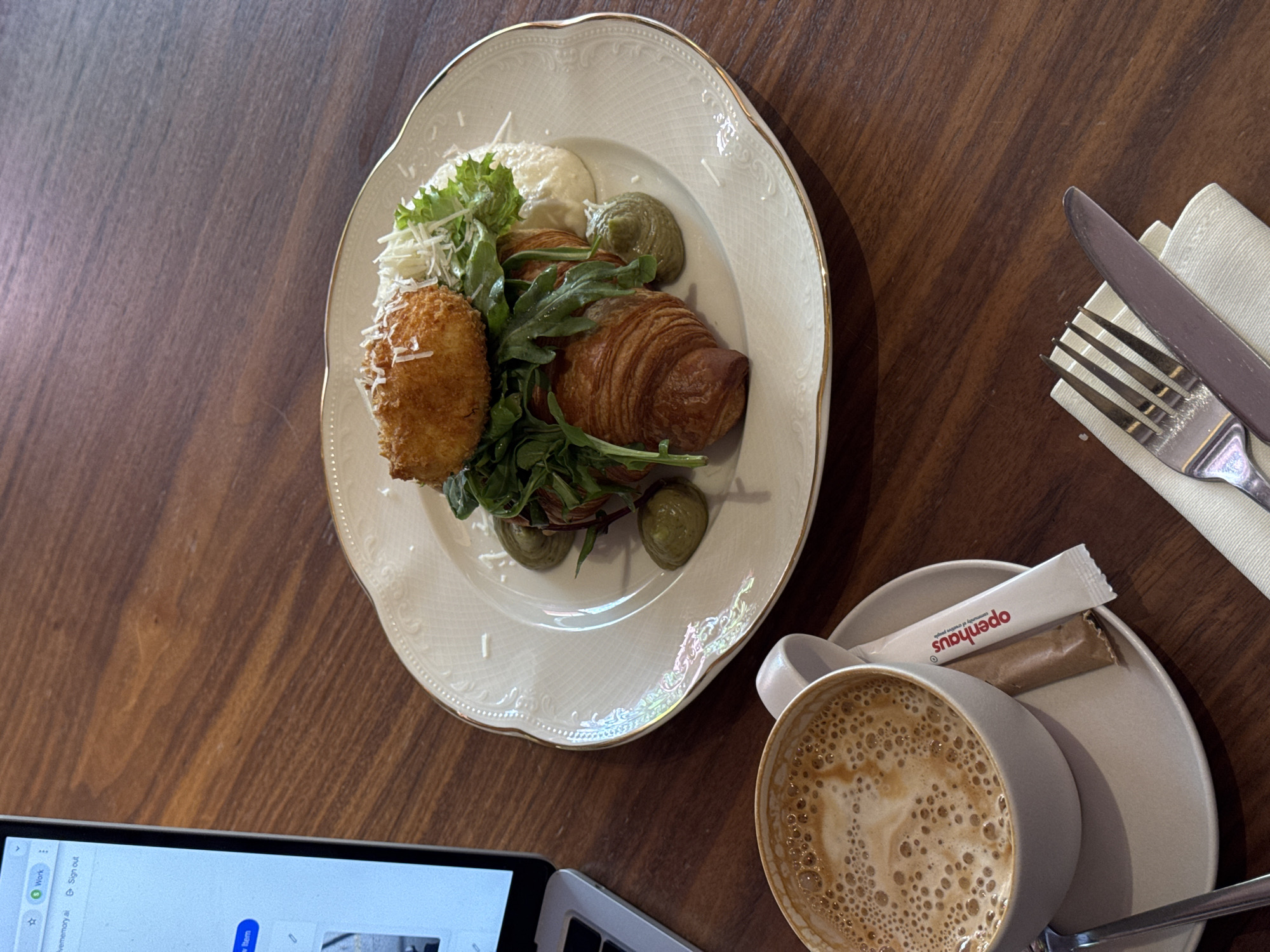 A plate of food consisting of a croquette, croissant, greens, shredded cheese, and small dollops of a green condiment is placed on a decorative white plate with gold trim. To the right of the plate is a fork and knife atop a white napkin. A mug of coffee and a saucer with a sugar packet and a packet of brown sugar are positioned on the right side of the frame. A laptop is visible on the lower left corner. The scene is set on a wooden table. The sugar packet is branded with the name "openhaus", which suggests the location is likely a café, bakery, or restaurant in Yerevan, Armenia where "openhaus" is a known establishment. No people are visible.