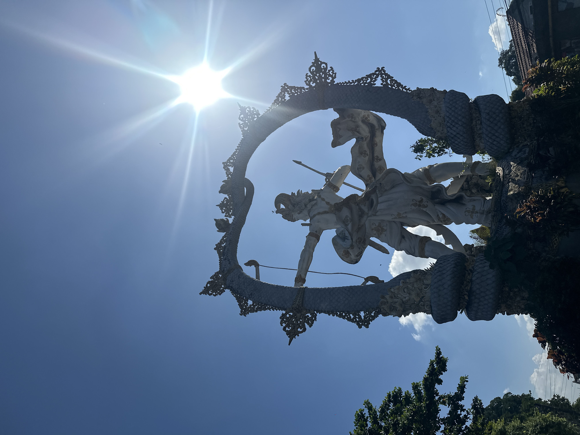 A low-angle, full-shot photograph captures a brightly lit, elaborate stone statue set against a vibrant blue sky on a sunny day. The statue depicts a figure in traditional attire, possibly mythological, holding a bow and arrow. The figure is framed by a large, intricately carved archway resembling a serpent, with decorative elements along its curves. The sun is prominent in the upper right quadrant of the image, emitting bright rays and lens flare. Green foliage and trees are visible to the left and bottom of the frame, and the roof of a building can be seen in the lower right. This scene is located in the Ubud District of Indonesia.