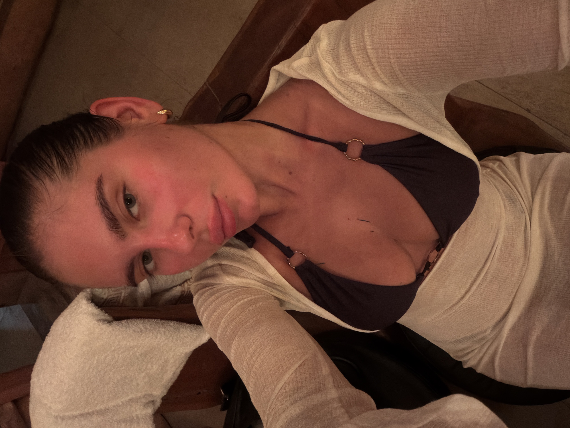 A young woman is captured in a close-up, angled selfie, looking directly into the camera with a neutral expression. Her dark hair is pulled back, and she has light-colored eyes. Her skin, particularly on her face and upper chest, appears flushed with a reddish tone. She is wearing a dark-colored bikini top with gold-toned O-rings on the straps, and over it, a sheer, loose-fitting, light cream or off-white long-sleeved cover-up. A small gold hoop earring is visible in her right ear.
The setting appears to be indoors or a sheltered area with dim, warm lighting, casting soft shadows. Hints of wooden surfaces, possibly paneling or furniture, are visible in the background, contributing to a cozy or intimate atmosphere. In the bottom left, a white fluffy towel is draped, and a dark strap or handle is partially visible near her left arm. The overall ambiance, combined with her attire, suggests a warm environment, possibly a spa, sauna, or a relaxed evening setting. Given the provided context, the location is Drokino, Russia. No visible text is present in the image.