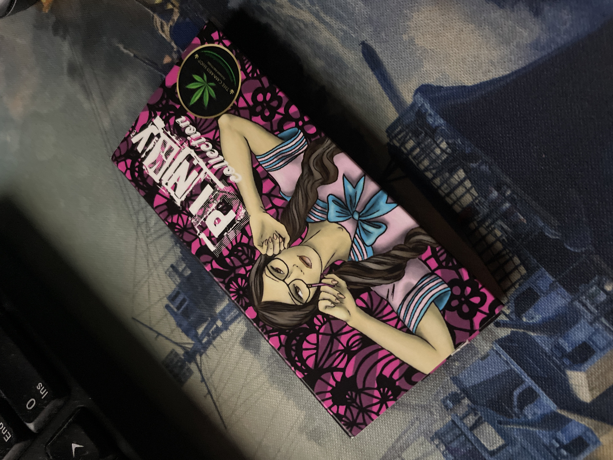 The image displays a rectangular box, oriented diagonally from the lower-left to the upper-right, resting on a textured, patterned surface. The box features a design dominated by hot pink and black geometric and floral patterns. Prominently displayed is the text 'PINKY collection' in white, stylized font. An illustration on the box depicts a light-skinned female figure with long dark hair, wearing round black glasses, a pink top with light blue striped sleeves, and a light blue bow at the neckline. Her left hand holds a slender pink object, possibly a pen, near her mouth, while her right hand adjusts her glasses. In the upper-right corner of the box, a circular black logo with a green cannabis leaf is visible, accompanied by the text 'THE CANNABIS SHOP' and 'ALL THINGS CANNABIS'.
The surface beneath the box is a fabric material, possibly a mousepad or desk mat, exhibiting a light grey background with dark blue/grey silhouettes of naval vessels and their superstructures. A black computer keyboard is partially visible in the upper-left corner of the frame, showing portions of the 'End', '0', and 'Ins' keys, along with a directional arrow key. The lighting suggests an overhead source, casting a subtle shadow from the box. No people are physically present in the scene, only the illustrated figure on the box. The scene is located in the city of Unknown, Unknown.