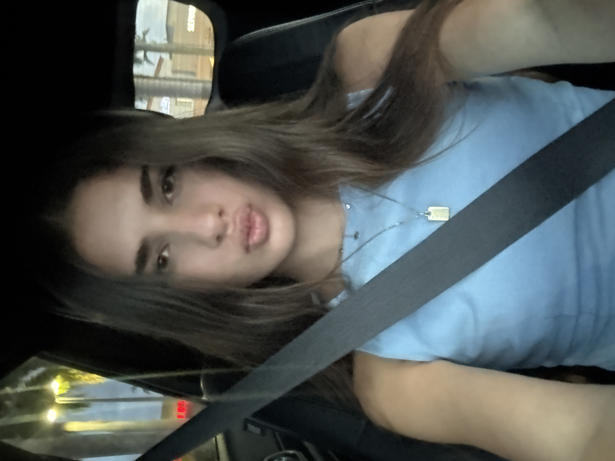 An image captures a young female in the driver's seat of a vehicle at night in Boca Raton, United States of America. The subject has long, dark brown hair, centrally parted, and wears a light blue sleeveless top. A dark gray seatbelt extends diagonally across her chest and right shoulder. She exhibits a "kiss face" expression with pursed lips. A silver-toned necklace is visible, featuring a small, circular charm and a rectangular, gold-toned pendant. The vehicle's interior appears dark. Outside the driver's side window, bright yellow streetlights, green foliage, and a red light are discernible. Through the passenger side window, a light-colored building, structures resembling palm trees, and a commercial sign with partially visible white text including "SERVI" on a dark background indicate a retail or service area.