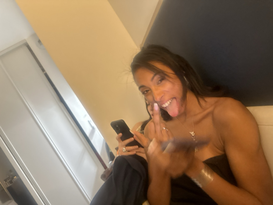 A woman smiles, sticks out her tongue, and raises her middle finger to the camera. She wears a strapless top and jewelry. Another woman sits behind her looking at a phone. The background includes neutral-colored walls and a white door. The image appears to be taken indoors with casual lighting.
