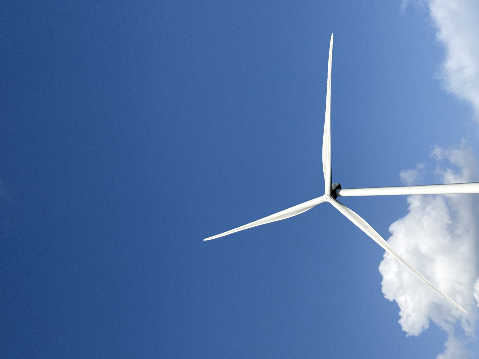 The image shows a close-up view of a wind turbine against a clear blue sky with a few white clouds near the bottom. The turbine's blades are white and slender, with one blade angled upwards and to the right, another angled downwards and to the left, and the third blade facing downwards and slightly obscured by the central column. The central column of the turbine is also white and appears to be a tall, cylindrical structure. The environment appears to be an open area, possibly a field or a coastal region, given the presence of clouds and the vastness of the sky. There are no people visible in the image, and the focus is entirely on the wind turbine and the sky.