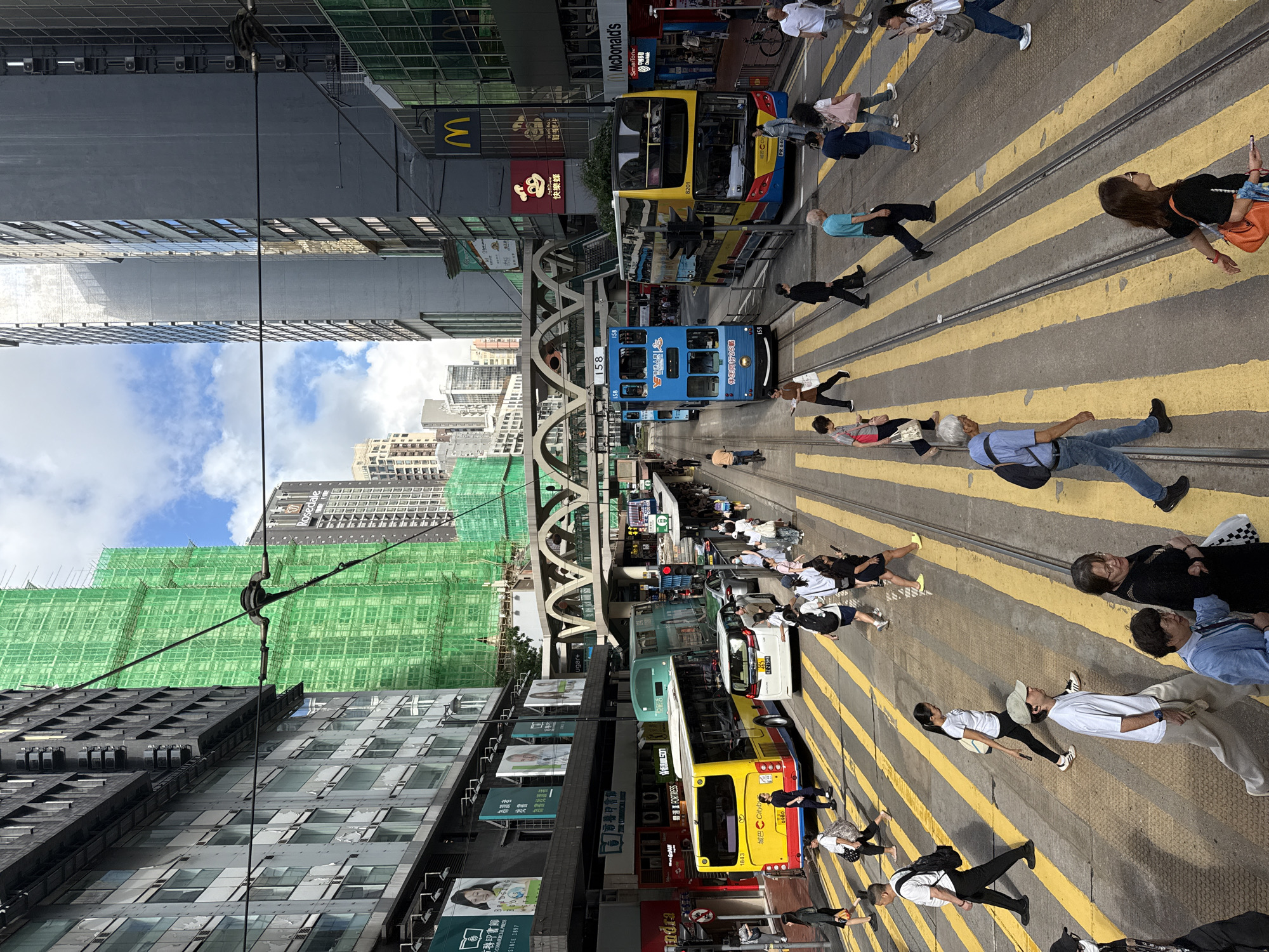 The image shows a street scene in Hong Kong, China, dominated by a tram line running through the center. People are walking on the pedestrian crossing over the tram tracks. Two trams, one blue and one yellow, are visible on the tracks, facing in opposite directions. Behind the trams are buildings under construction, covered in green scaffolding netting. Buildings line both sides of the street, and a McDonald's restaurant sign can be seen. Pedestrians are walking along the crosswalks, crossing the tracks. A raised walkway spans over the tram tracks. The sky is visible between buildings, showing some clouds.
