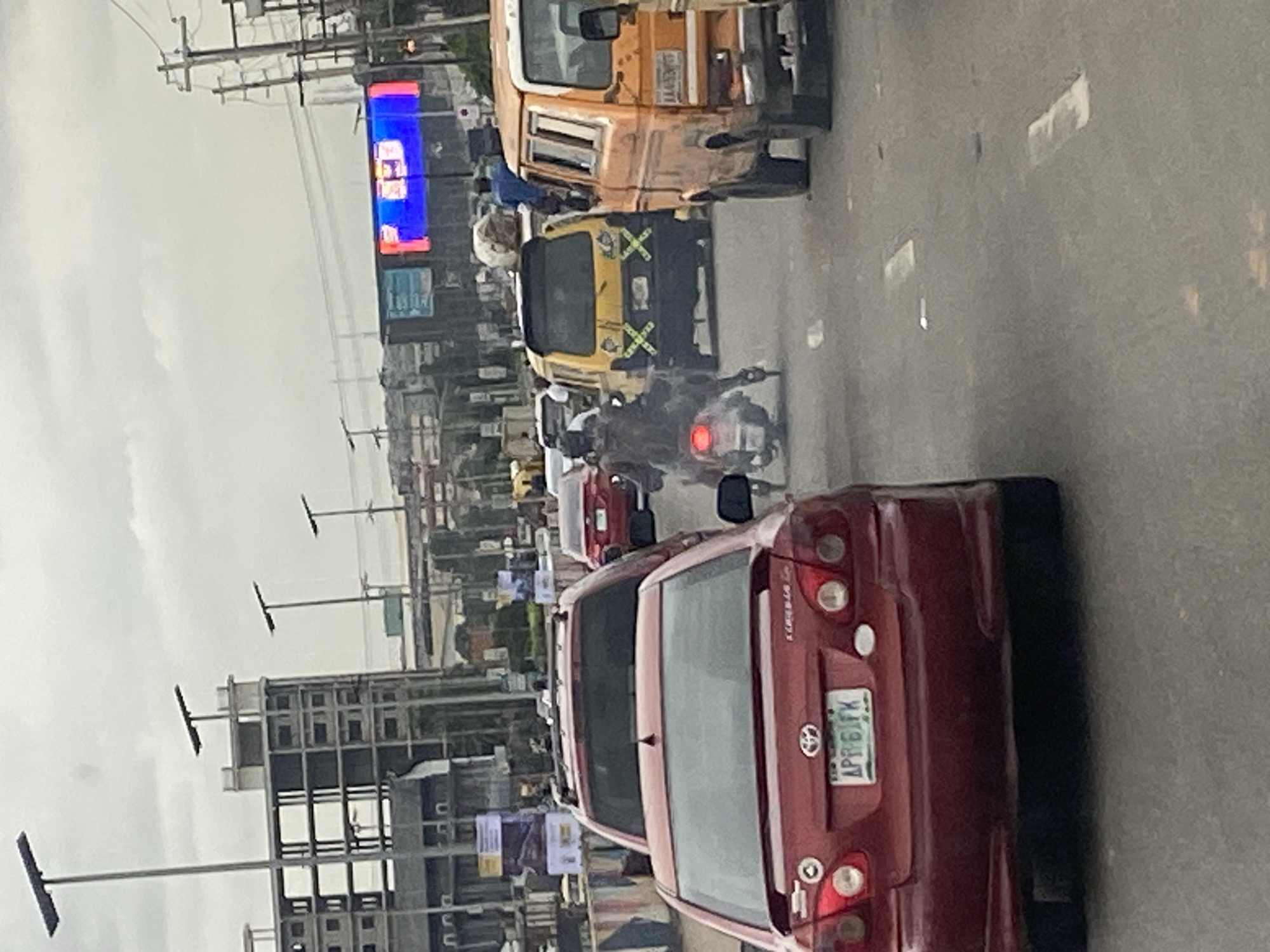 This is a forensic, matter-of-fact description of a traffic scene in Ikeja, Nigeria. The image depicts a busy roadway with several vehicles, including a red Toyota Corolla in the foreground. The Corolla has a license plate that reads "APR 61FK". Behind it, a motorcycle rider wearing a helmet is visible. Further down the road, a yellow mini-bus with yellow and black X-shaped markings on its rear is present, along with an orange van. In the background, construction is underway on a large building, and several streetlights are visible. A large digital billboard displaying blue and red content is also noticeable. The sky is overcast, suggesting a cloudy day. The scene captures a moment of typical urban traffic congestion in Ikeja.