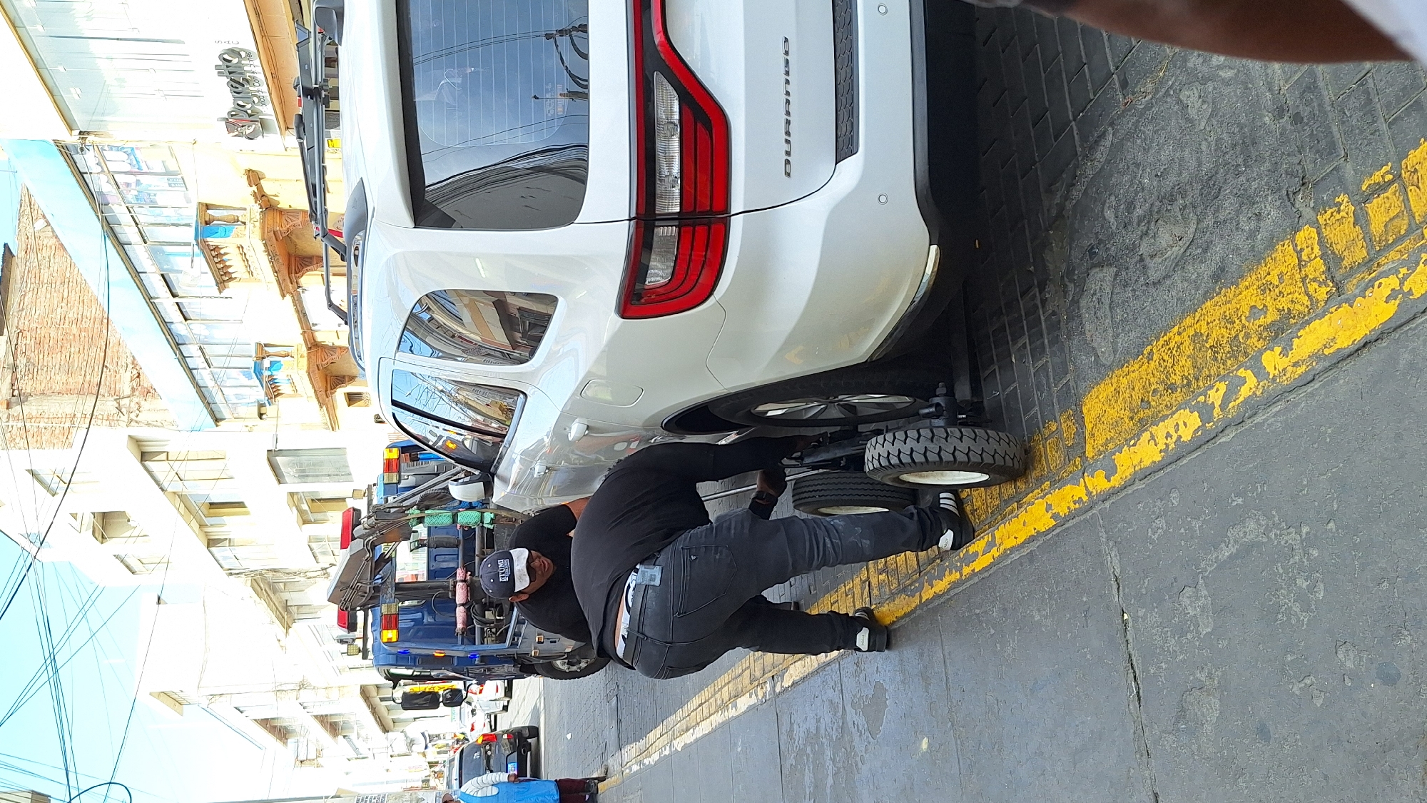 The image displays an outdoor urban street scene during bright daylight. A man, dressed in a black t-shirt, black jeans, and black and white sneakers, is actively working on the rear underside of a white SUV. He wears a black baseball cap with white text, possibly "BEYOND". A small, wheeled device with two tires is positioned beneath the SUV's rear axle, suggesting the vehicle is either being lifted, prepared for towing, or undergoing maintenance. The SUV is identifiable as a Dodge Durango, with the word "DURANGO" visible on its tailgate.
The man is crouched low, appearing focused on his task. The street features worn grey pavement with distinct yellow painted lines. In the background, a blue pickup truck and other vehicles are visible along the street. The buildings lining the street exhibit varied architecture, including one with exposed brickwork and others with multiple windows and balconies. Overhead utility wires are visible against the clear sky. The scene depicts a routine street activity, likely a vehicle recovery or repair operation.