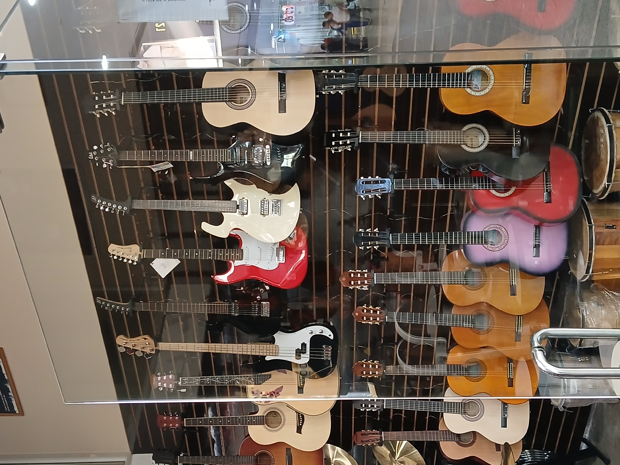 The image depicts a display of various guitars and musical instruments, likely in a store or shop in Guayana City, Venezuela. A range of guitars, including electric, acoustic, and classical models, are visible. Some drums are also present within the display. The objects are arranged on a wall or behind a glass enclosure. The reflection on the glass indicates an indoor setting, possibly a commercial space.
