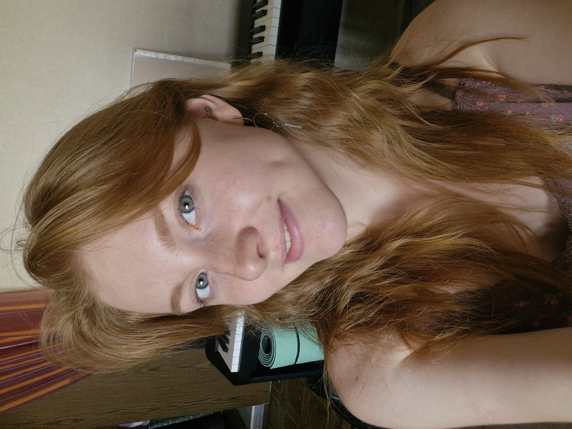 A portrait of a young woman with auburn hair and fair skin. She has blue eyes, freckles, and a faint smile. She is wearing a floral-patterned top and has earrings and a piercing in her ear. She is positioned at an angle to the camera. Behind her is a keyboard, a rolled-up yoga mat, and a wooden surface. The location is not identifiable as Yekaterinburg, Russia, based on the image provided.