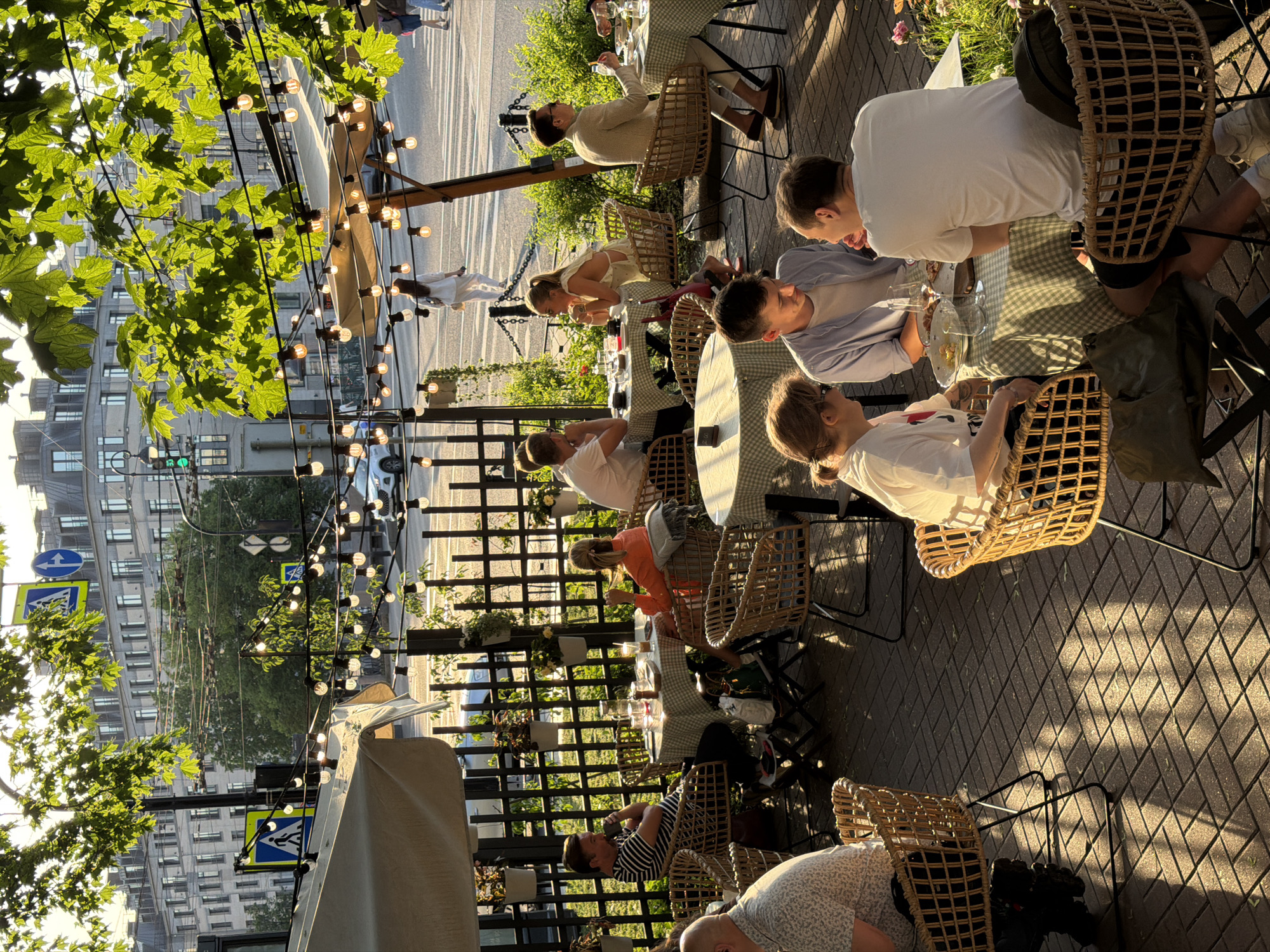 The image depicts an outdoor seating area of a restaurant or cafe on a street in Saint Petersburg, Russia, bathed in the warm, golden light of late afternoon or early evening. Tall, leafy trees with dappled sunlight filtering through their branches provide shade, and a string of illuminated lights stretches across the scene. In the background, a multi-story building with a grand architectural style, characteristic of Saint Petersburg, stands prominently. The street itself appears to be a busy thoroughfare, with a white car visible and pedestrian crossings marked on the pavement.
The foreground and midground are occupied by several tables and chairs where patrons are dining and socializing. The furniture is made of woven rattan or wicker, with a rustic yet chic aesthetic. The tables are covered with checkered tablecloths in shades of green and white.
Several groups of people are visible:
* A man in a striped t-shirt is seated at a table, holding a cup to his lips.
* A woman in an orange top is seated opposite him, looking down at her table.
* A man in a light-colored suit is seated at another table, holding a fork and looking towards his companions.
* A woman in a white top, with blonde hair tied back, is seated at a table, her hands clasped.
* A man in a light blue shirt is engaged in conversation with a woman in a white t-shirt, both seated at a table.
* Another man in a white t-shirt is also seated at this table, his back to the camera.
* A man in a patterned shirt is visible at the far left, his head turned away.
The overall atmosphere is relaxed and convivial, suggesting a pleasant dining experience in a charming urban setting. The dappled sunlight and shadows cast on the brick-paved ground add a dynamic visual quality to the scene.