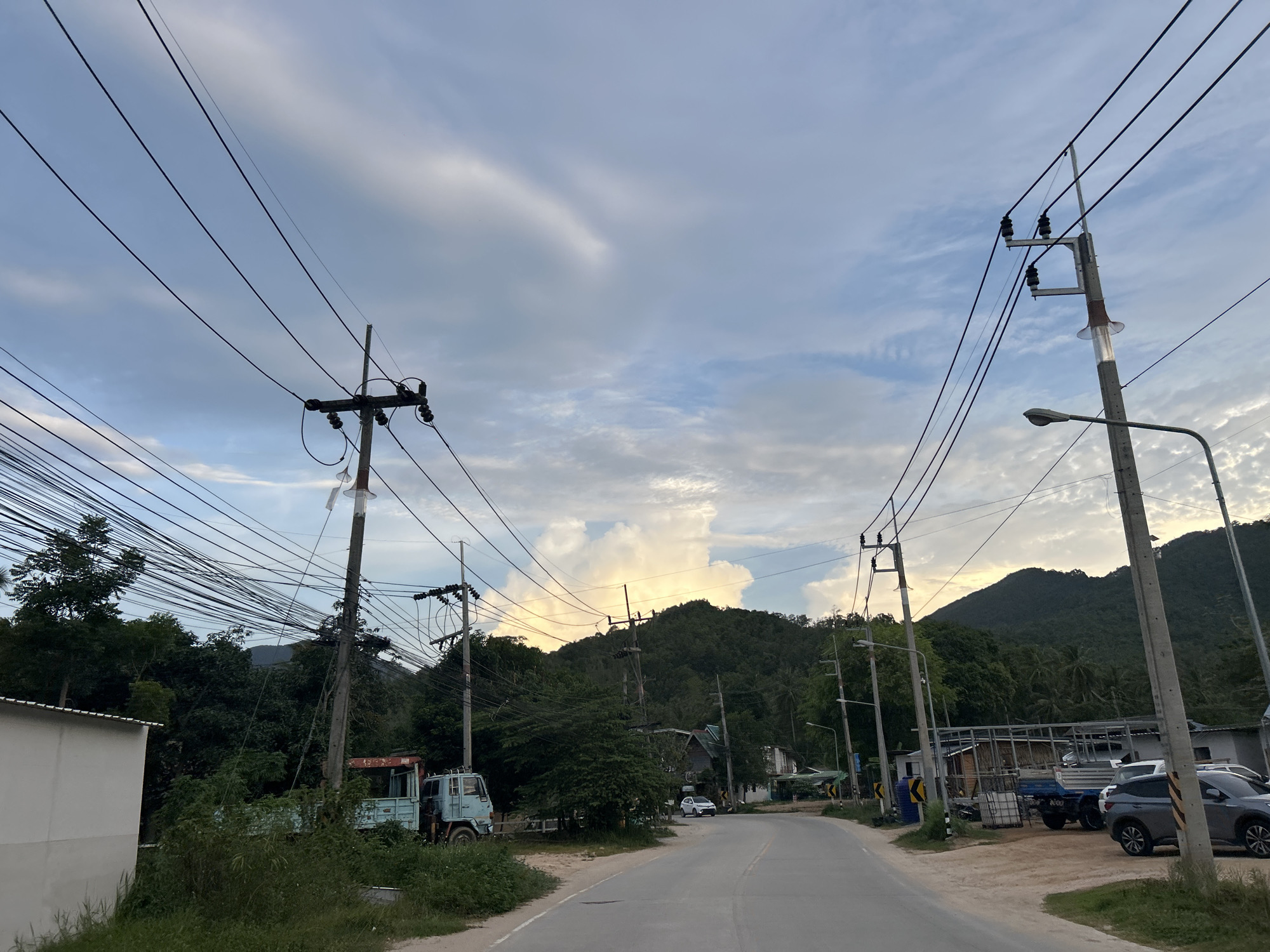 This image depicts a road in the city of Ko Pha-ngan District, Thailand, under a cloudy sky. The road stretches into the distance, flanked by vegetation and buildings. Several utility poles with numerous electrical wires are prominent in the foreground and midground, with one pole also featuring a street lamp. A light blue truck is parked on the left side of the road, partially obscured by foliage. Further down the road, several vehicles are visible, including a dark gray SUV parked on the right side. In the background, a lush, tree-covered mountain range dominates the horizon. The lighting suggests it is either early morning or late afternoon.