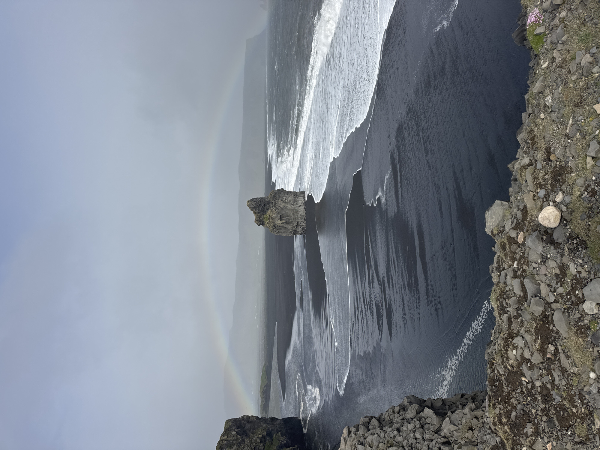 This photograph captures a high-angle view of Reynisfjara black sand beach near Vik, Iceland. The scene includes a segment of a rainbow arching across the sky on the left, a basalt sea stack in the ocean, and waves crashing onto the black sand. The right side of the frame shows a rocky cliff edge with vegetation.
