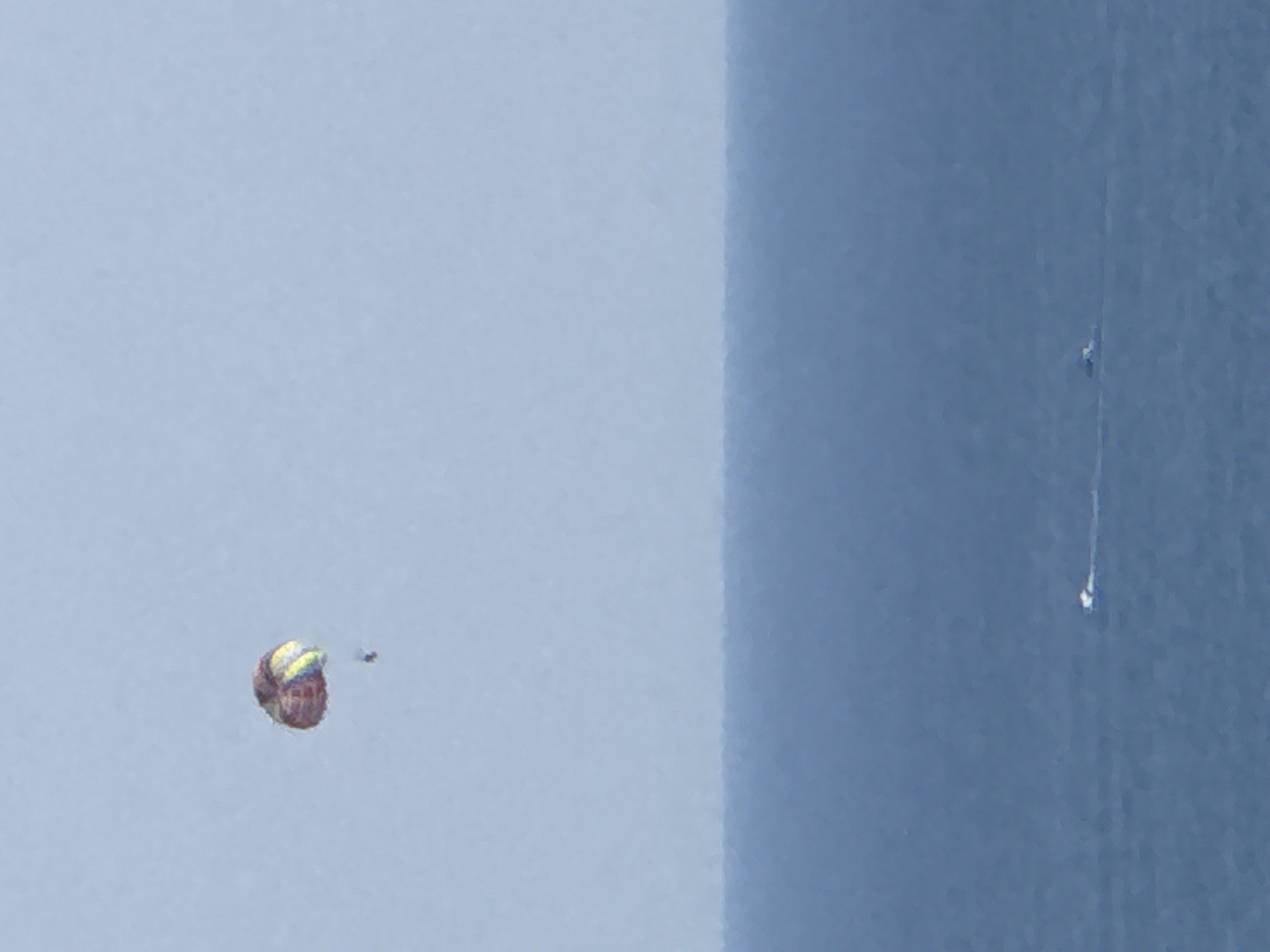 The image shows a parasailer in the sky above a body of water. The parasail is multicolored, with red, yellow, and blue sections. A person is visible hanging from the parasail. In the water below, there are two small boats leaving white wakes. The horizon is visible, separating the pale blue sky from the dark blue water. The scene appears to be a recreational activity on a clear day. The location is stated to be Boreti, Montenegro, suggesting a coastal or lake setting.