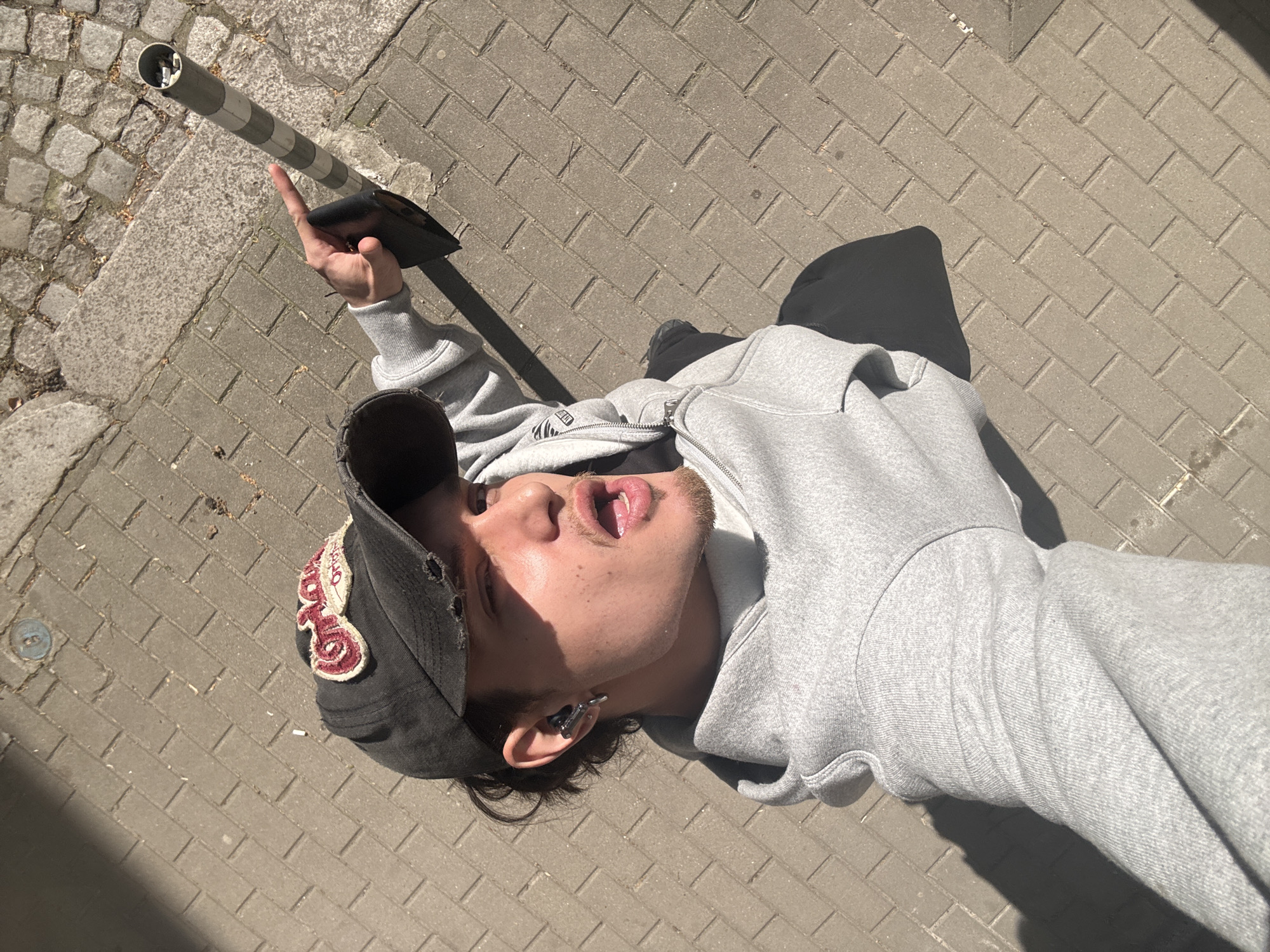 The media features a young adult male lying on a paved sidewalk or street, captured from an overhead perspective under bright sunlight. The scene is set in an urban environment, identified as Sofia, Bulgaria.
The subject is wearing a gray zippered hoodie and a black baseball cap with a red embroidered patch and distressed details. He has light facial hair and an ear cuff or earring on his left ear. He is positioned horizontally in the frame, looking upwards with his mouth open and tongue visible, possibly indicating a casual, surprised, or relaxed state. His right hand holds a black smartphone, pointing towards a striped metal pole (likely a bollard or signpost) in the upper left portion of the image.
The ground is predominantly composed of gray rectangular pavers laid in a herringbone pattern, with a section of cobblestones visible to the upper left. Distinct, elongated shadows are cast by the person and the pole, suggesting a sunny day, likely mid-morning or afternoon. Notable details on the ground include a small, white cigarette butt near the cap and a circular metal utility plate embedded in the pavement. The red patch on the cap appears to have stylized lettering, but no specific text is clearly legible.