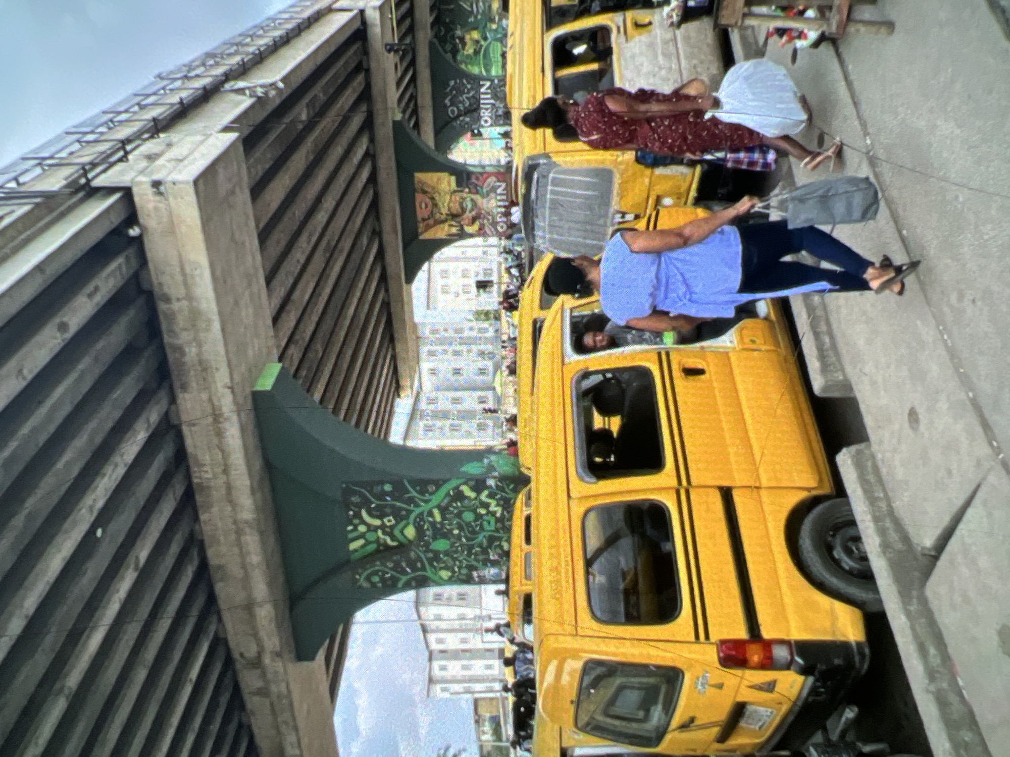 The image depicts a bustling scene in Lagos, Nigeria, under a large concrete overpass. Several yellow minibuses, commonly known as danfos, are parked along a sidewalk. The danfos are bright yellow with black stripes and appear to be in use for public transportation.
In the foreground, two women are walking on the sidewalk. The woman on the left wears a light blue top and dark pants, carrying a gray bag. The woman on the right is wearing a patterned red dress and also carrying a white bag. Both are wearing sandals and appear to be moving away from the viewer.
The overpass structure is prominent, with its concrete beams and supporting pillars. The pillars are painted green and decorated with intricate murals of foliage. The sky above is overcast. In the background, multi-story buildings are visible, suggesting an urban environment. The overall impression is one of everyday life and activity in a busy city.