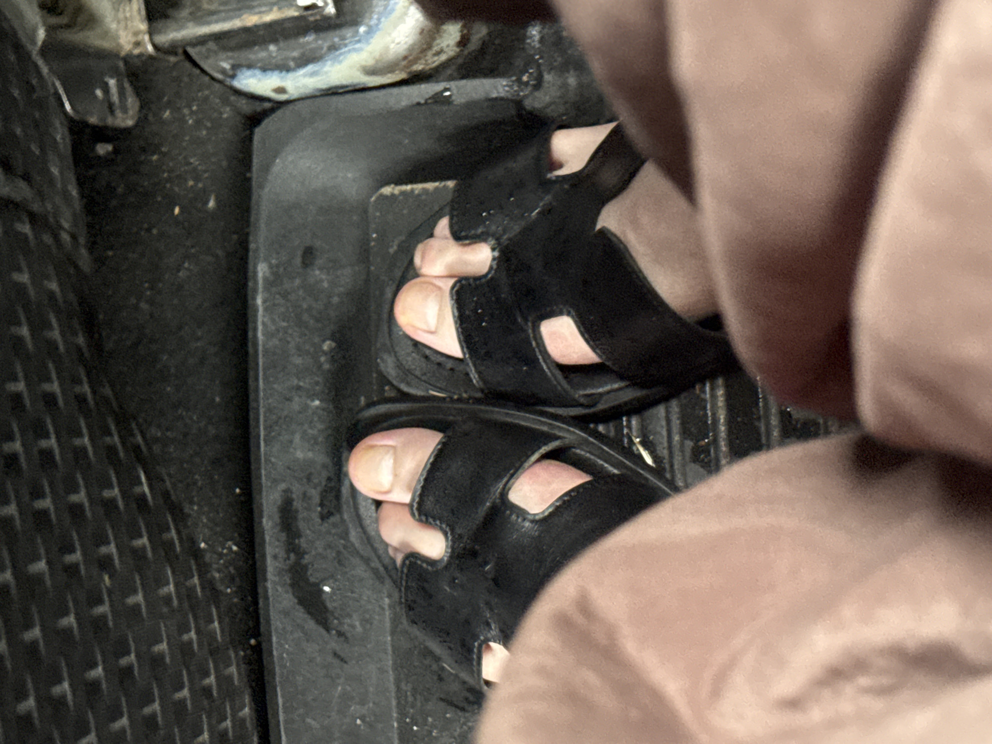 The image shows a close-up view of a person's feet wearing black sandals, resting on a textured floor mat inside a vehicle. The feet are positioned on the floor near the pedals. The environment appears to be the interior of a car, likely in the driver's or passenger's footwell. The lighting suggests an indoor setting or possibly a dimly lit area. There is no visible indication of the specific location within the city of Tiraspol, Moldova, or any activity other than the person resting their feet.