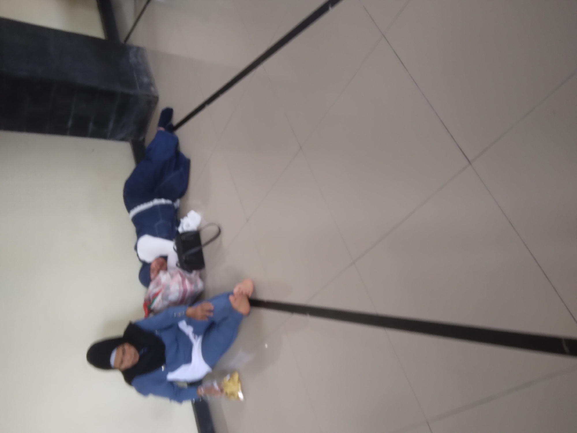 Two individuals are observed on the tiled floor, positioned against a light-colored wall. The person on the left is seated upright, wearing a blue and white hijab and a blue garment, with their legs extended. They are holding a yellow packet in their left hand. Beside them lies a large bag filled with items. To the right, a second person is lying down on their side, facing away from the camera, wearing a dark blue hijab and a blue garment. A black handbag is placed near them. The environment appears to be an indoor waiting area, indicated by the tiled floor and the wall. The lighting is subdued, suggesting it might be indoors during daylight hours. The scene conveys a sense of rest or waiting.