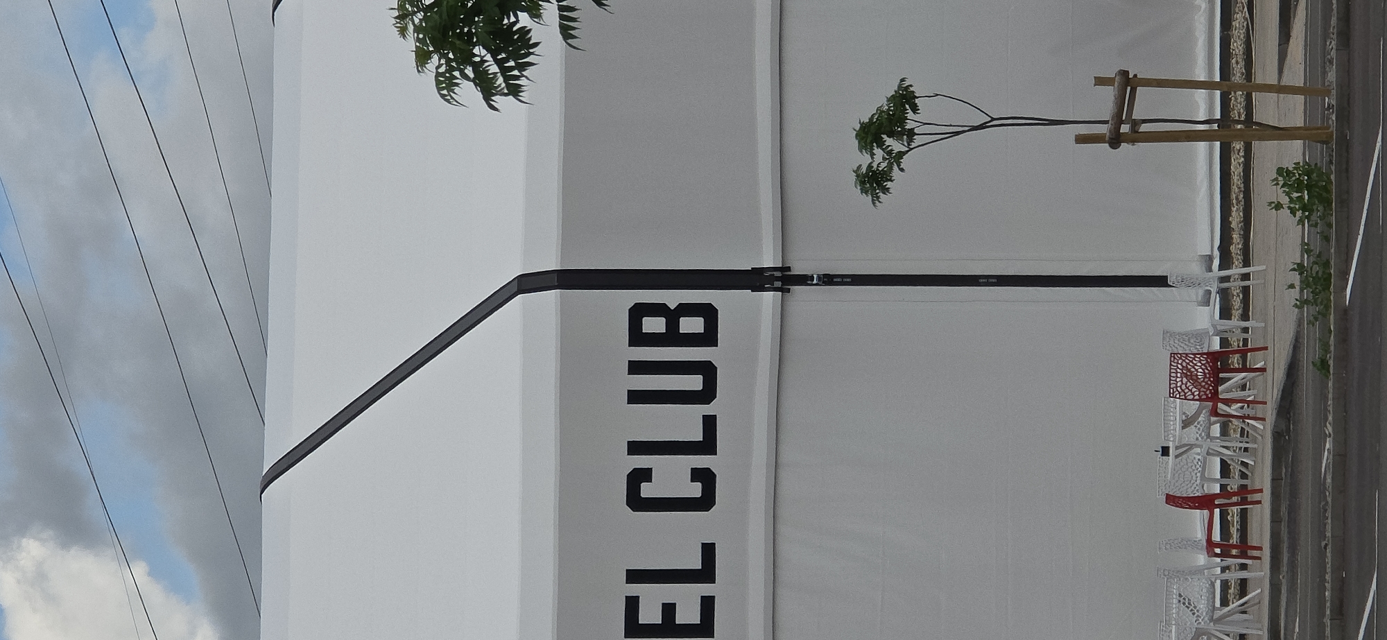 The image depicts an outdoor scene dominated by a large, white, tent-like structure or building. A prominent black vertical band runs down the side of this structure, featuring bold black letters that read "EL CLUB" when viewed upright. Another black line forms a diagonal detail on the structure's surface.
To the right of the structure, an outdoor seating area is visible. It comprises several modern-looking chairs, primarily white, with at least two red chairs scattered among them. Some chairs have decorative, perforated backrests and seats. Adjacent to the chairs and the structure, there is a paved ground surface, and a simple wooden barrier or fence runs along it. Small green bushes and foliage are visible near the ground and also higher up, with a slender tree trunk and branches reaching across the white structure in the upper right.
The sky in the upper left corner of the image is partly cloudy, with several dark power lines stretching horizontally across it. The overall lighting suggests it is daytime. No people are visible, and there is no specific activity taking place, though the arrangement of chairs implies a potential waiting or dining area, possibly for a club or establishment indicated by the visible text.