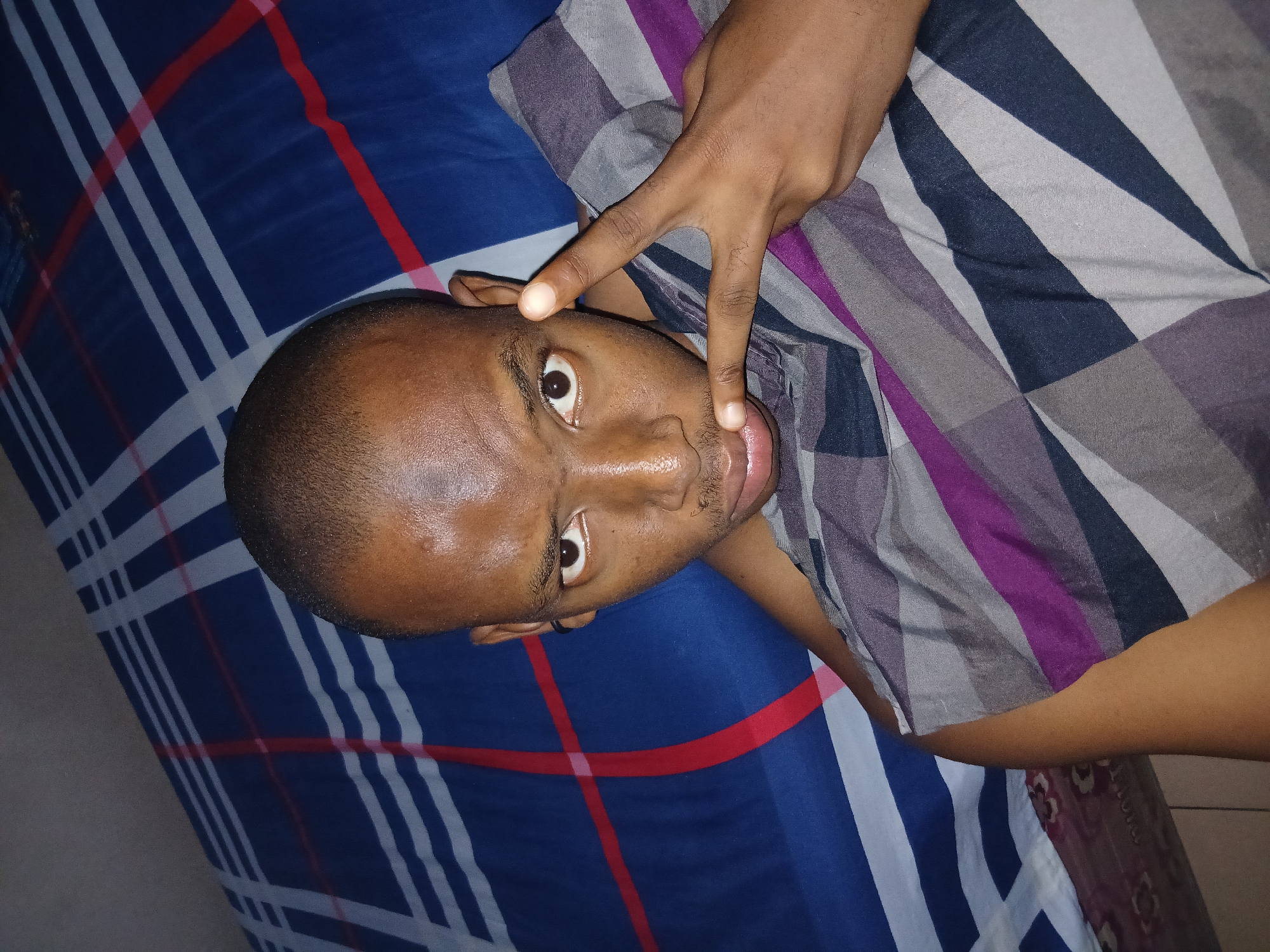 A young Black male is captured in a close-up, over-the-shoulder selfie, lying down. He has a shaved head, dark skin, and is looking directly at the camera with a neutral expression. A small mustache is visible above his upper lip. His right hand is raised, forming a peace sign gesture in front of his face. He is partially covered by a blanket with a geometric pattern of dark blue, purple, grey, and white diagonal stripes. The background consists of a bedspread with a blue, white, and red striped pattern, suggesting an indoor setting. The lighting appears to be artificial and overhead. The image does not contain any specific landmarks or details that would allow for precise identification of a location within Lagos, Nigeria; it is a generic domestic interior scene.
