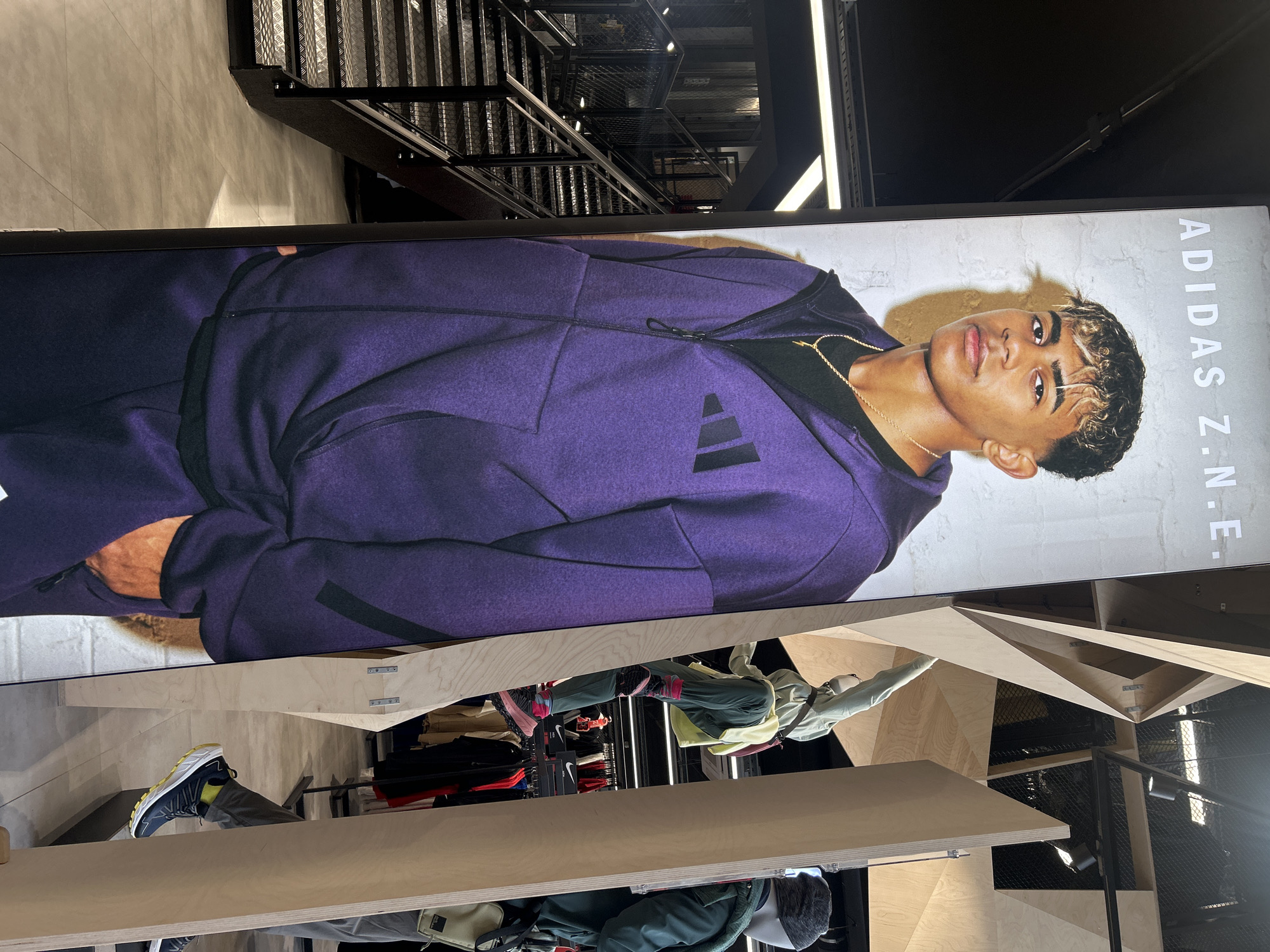 The image shows a retail space, possibly an Adidas store, based on the large poster featuring an Adidas Z.N.E. advertisement with a person wearing a purple Adidas jacket. The poster is mounted vertically against a textured, light-colored wall. A multi-level, geometric wooden display structure is in the foreground. The display holds clothing items and mannequins dressed in sportswear. Further in the background, there's a staircase with a metal railing. The scene is illuminated by overhead lighting.