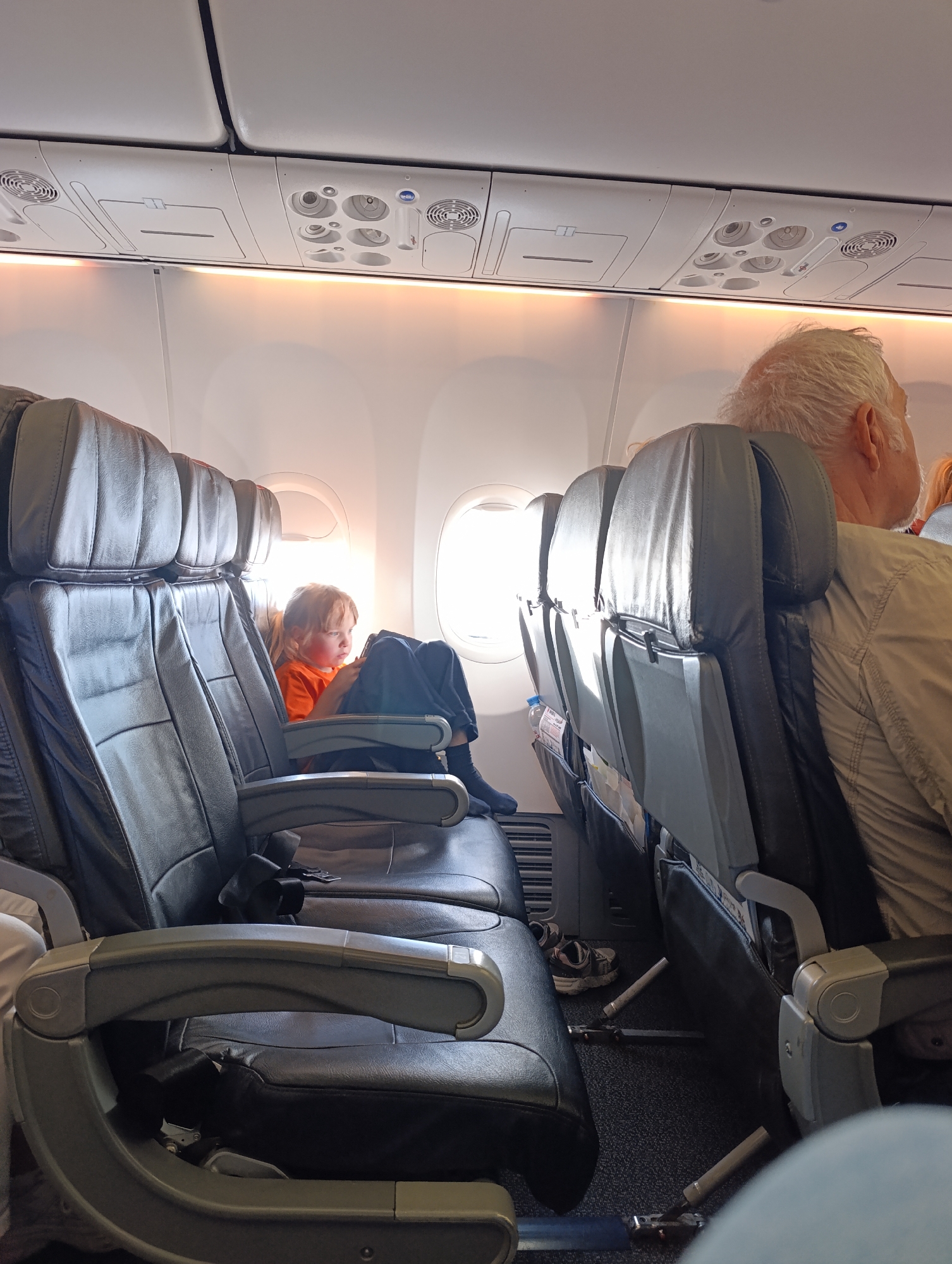 The image depicts the interior of an airplane cabin, specifically looking forward and slightly to the right from a passenger's perspective. In the foreground and to the left, dark leather airplane seats are visible, one of which is occupied by a young child with blonde hair wearing an orange shirt. The child is looking at a handheld device. To the right of the child's seat, another row of airplane seats is visible, with the back of an elderly man with gray hair, wearing a light-colored shirt, in the closest seat. The man's head is turned away from the camera, looking towards the right. Sunlight streams in from the windows, illuminating the cabin and casting a warm glow. Above the seats, the airplane's overhead compartment and air vents are visible. The floor of the cabin is carpeted. No specific location in the city of Staroe Mart'yanovo, Russia, can be determined from this image.
