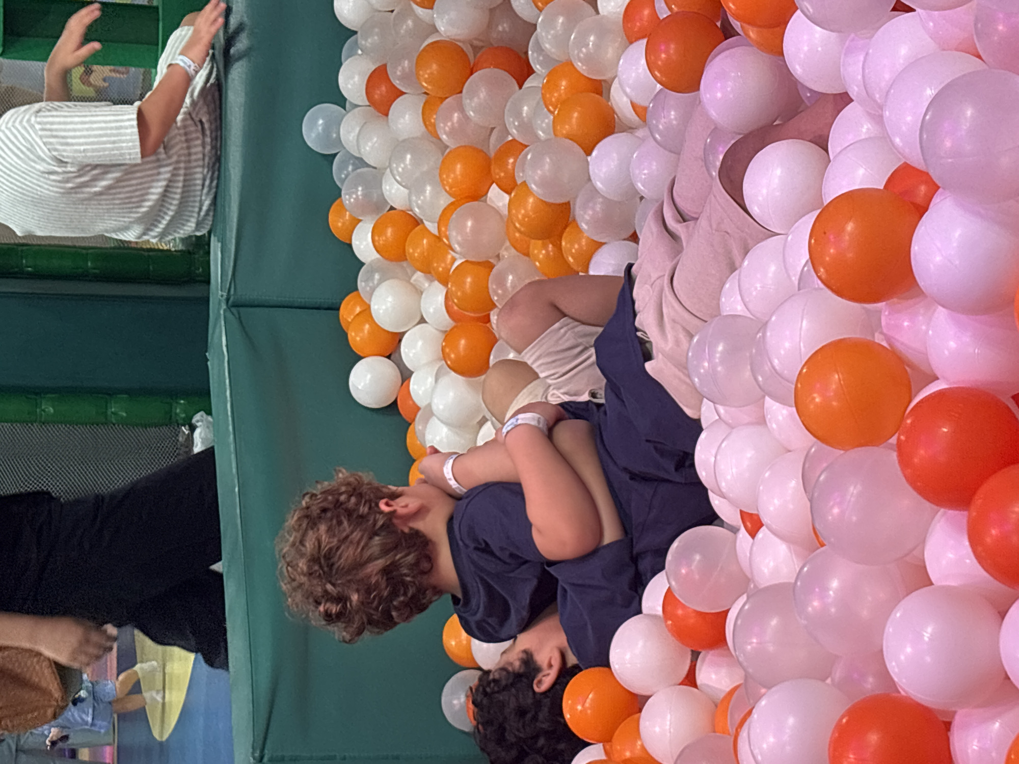 The image shows two young boys embracing in a ball pit filled with white, pink, and orange balls. The boy in the front has dark curly hair and is wearing a navy blue t-shirt and pants. He is lying on his side, with his head on the other boy's lap. The second boy, who is embracing the first, has lighter curly hair and is wearing a striped t-shirt and shorts. Both boys have white wristbands on their wrists. The ball pit appears to be indoors, with a green padded wall visible in the background. The environment suggests a children's play area. There are no visible signs or landmarks indicating a specific location within Dubai, United Arab Emirates.