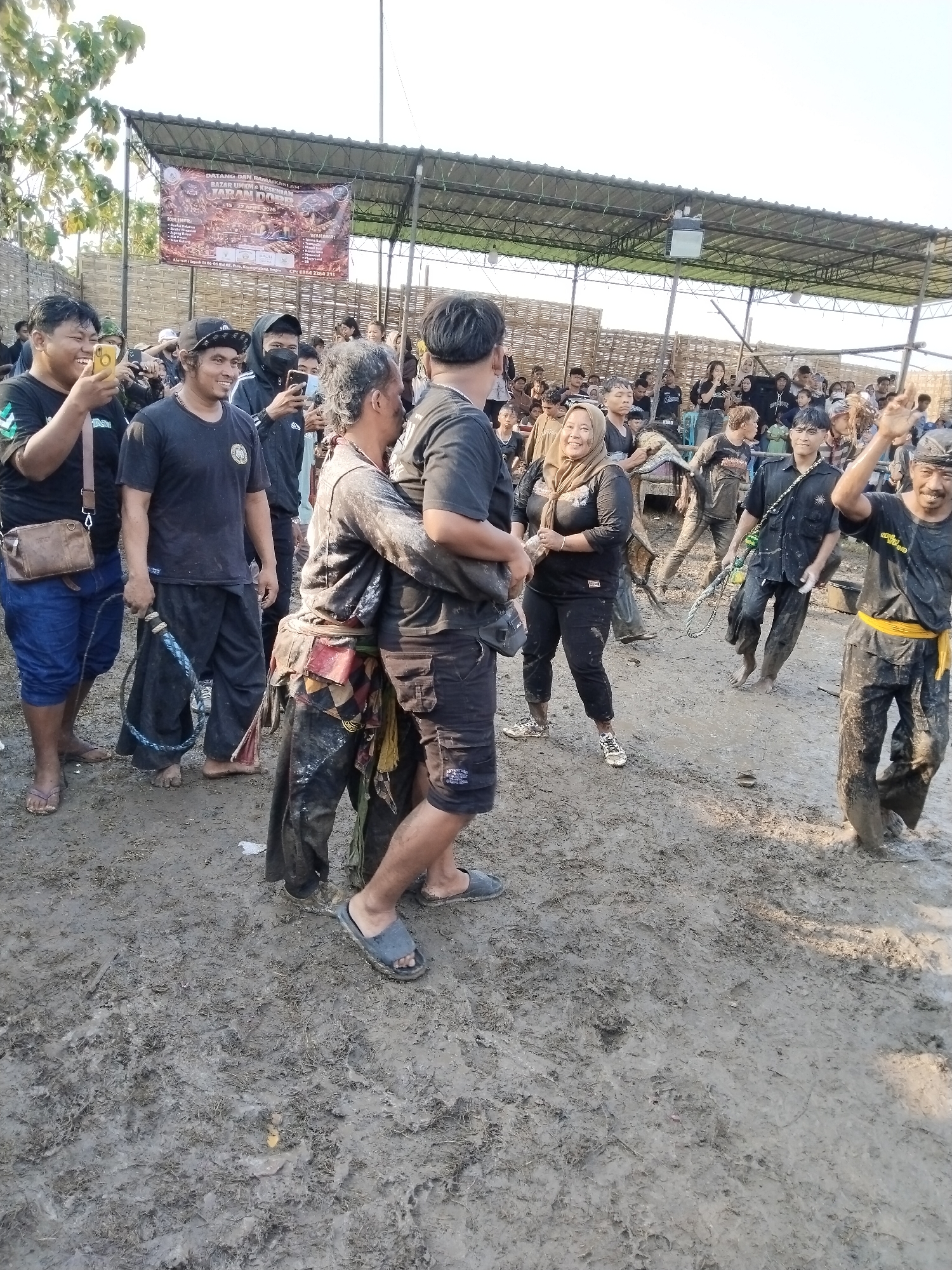 The image captures a lively outdoor cultural event in Kedawung, Indonesia, under a bright, clear sky. A crowd of people, mostly adults and young adults, are gathered, with many participants covered in mud. The muddy ground is a prominent feature of the scene, with many individuals barefoot or wearing sandals.
In the foreground, a central pair of men are caked in mud, with one carrying the other piggyback, both displaying expressions of amusement. To their left, a man holds a whip-like object, while another nearby grins and looks at his phone. On the right, a man with a yellow sash, also covered in mud, waves his arm energetically and smiles broadly. Another muddy man in the mid-ground holds a rope or chain. A woman wearing a hijab, visibly splattered with mud, stands nearby, smiling, indicating her active participation in the event.
Spectators are visible in the background, some observing from behind a bamboo fence or on elevated structures beneath a corrugated roof canopy. Several people are seen using their phones to record the event. The overall atmosphere appears joyful and engaging, with smiles and laughter evident among the participants.
A large banner hangs above the crowd, providing event details. The text on the banner reads: "DATANG DAN RAMAIKAN BAZAR UMKM & KESENIAN JARAN DORR" (Come and liven up the MSME Bazaar & Jaran Dorr Art) with dates "15 - 22 APRIL 2026." It also mentions "KULINER:" (Culinary), an address "Segush 106-06 w eu, fure, Kedawung," and a contact number "CP: 0864 2354."
